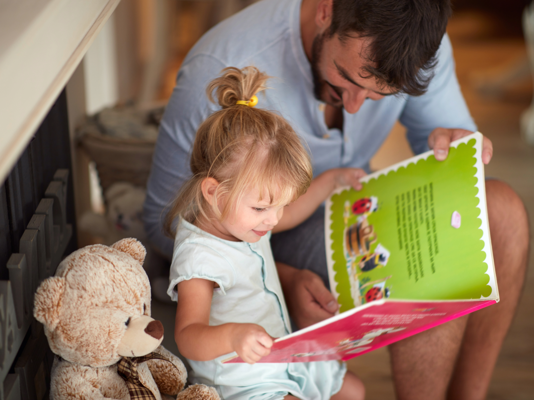 Vater und kleine Tochter schauen gemeinsam in ein Bilderbuch; daneben sitzt ein Teddybär.
