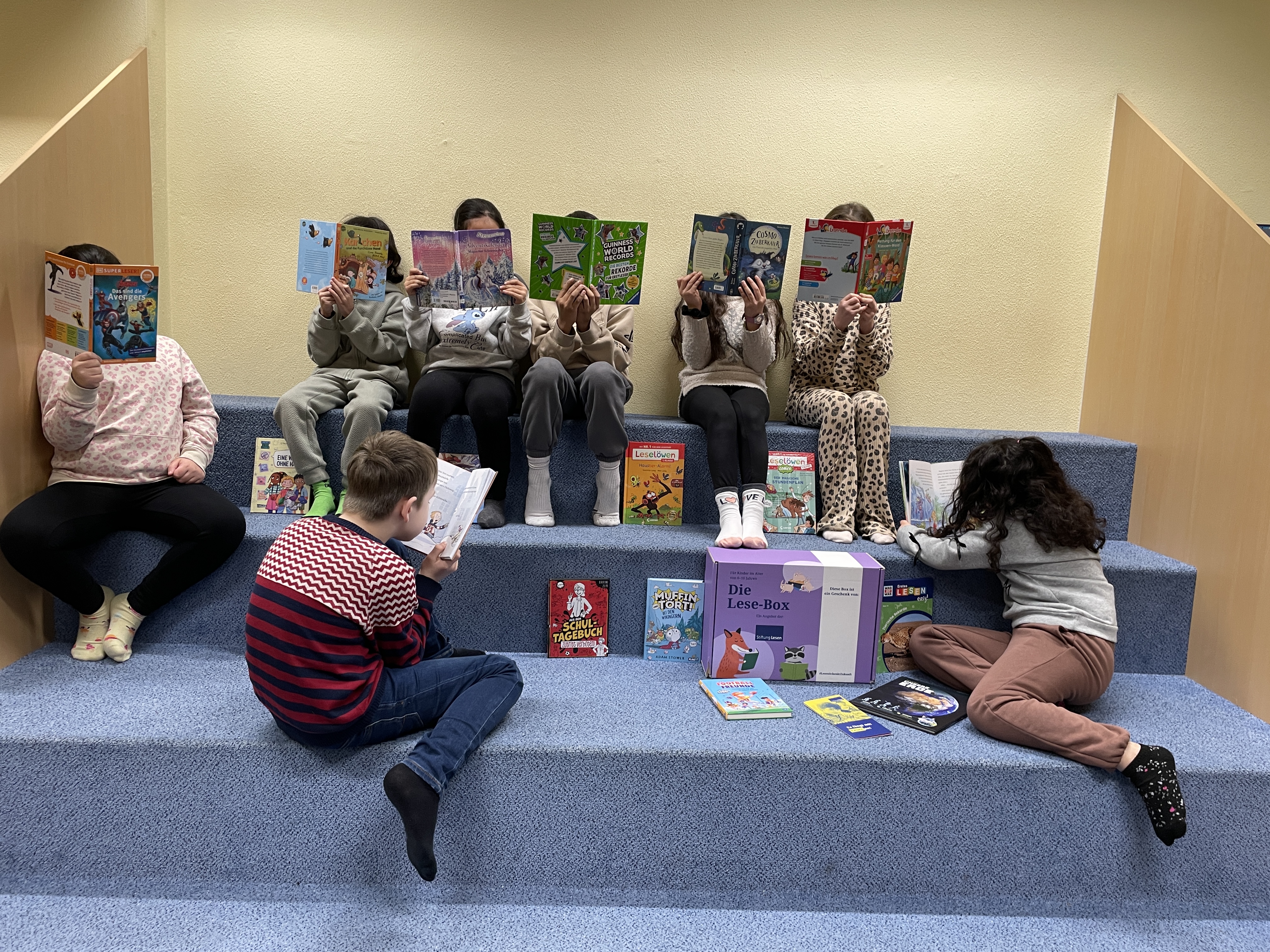 Kinder sitzen auf einer stufenförmigen Leseecke und lesen konzentriert in Büchern; vor ihnen liegt eine „Die Lese-Box“ mit weiteren Kinderbüchern.