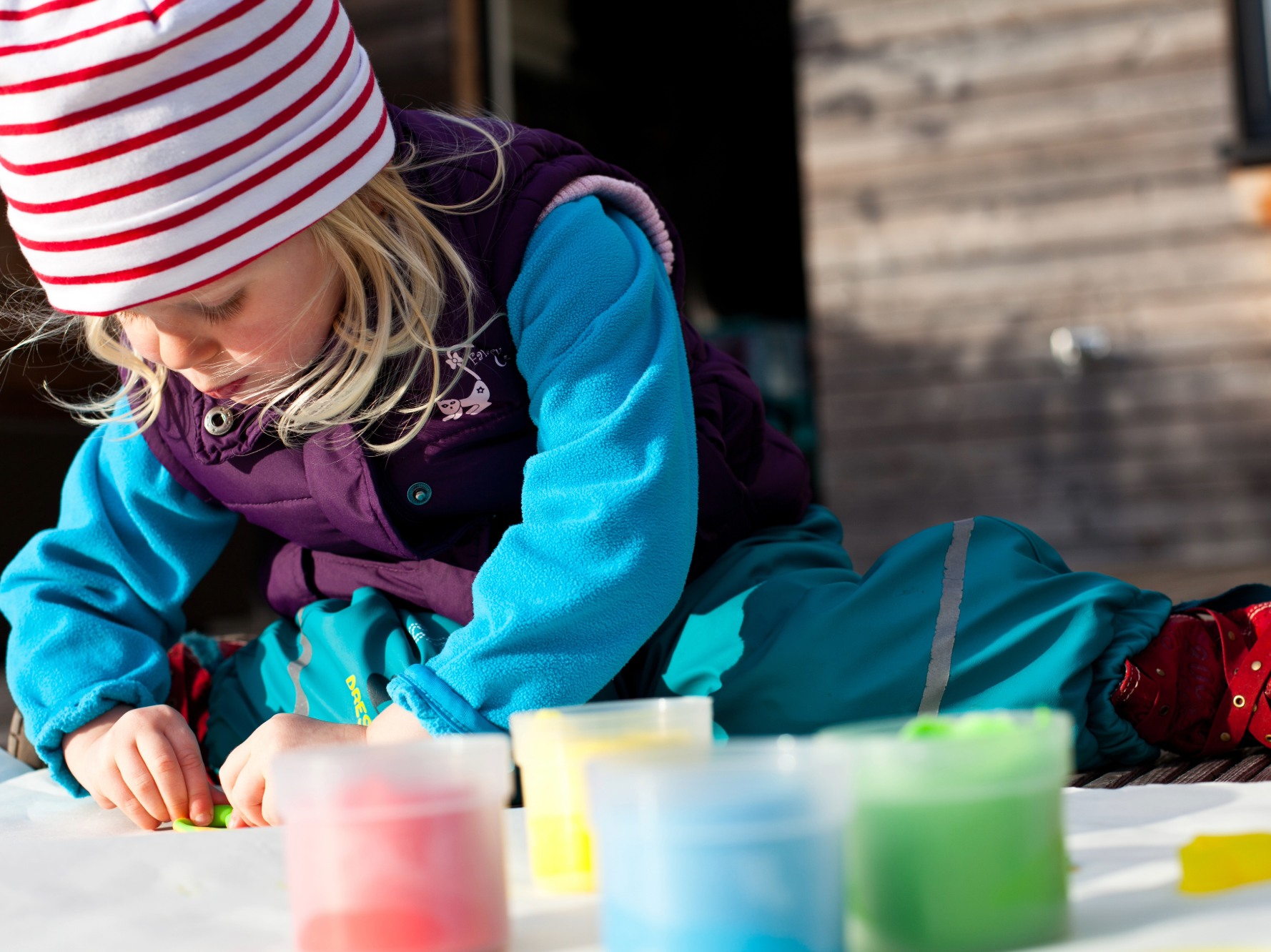 Kind in bunter Outdoor-Kleidung bastelt konzentriert an einem Tisch mit Farben im Vordergrund.