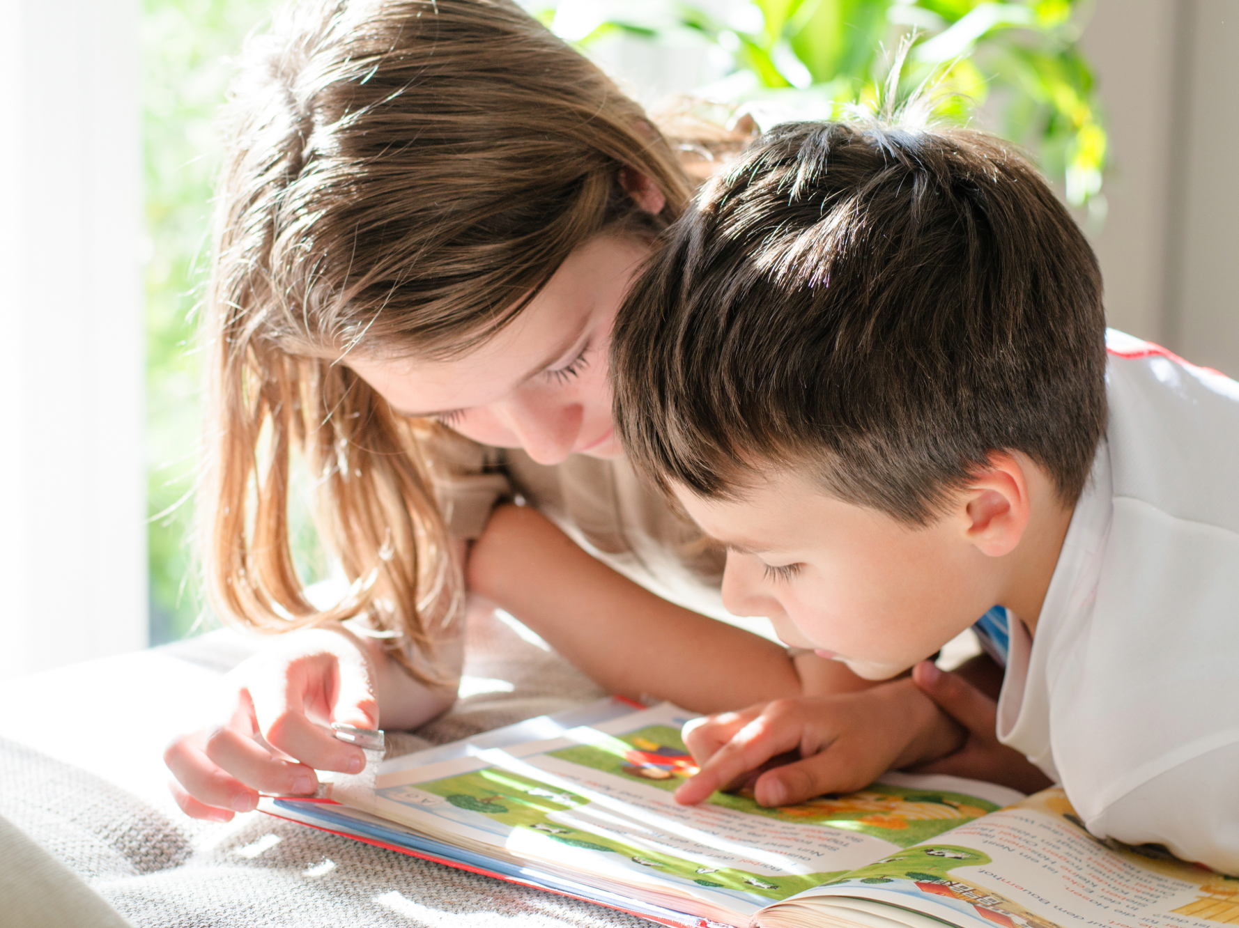 Zwei Schulkinder liegen nebeneinander und betrachten gemeinsam aufmerksam ein aufgeschlagenes Buch. Eines der Kinder zeigt mit einem Stift auf eine Seite, während das andere interessiert mitliest.