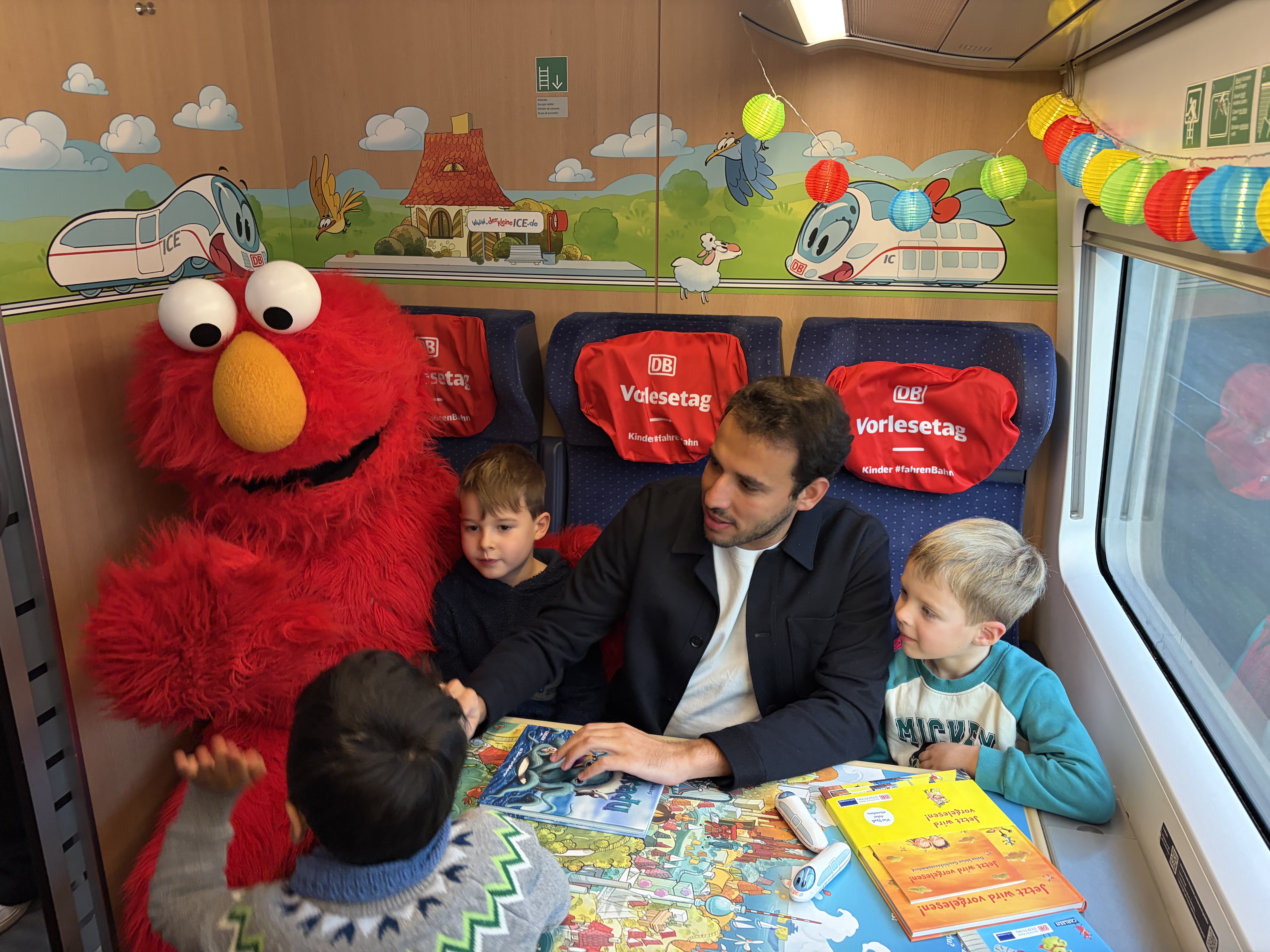 Ein Mann (Sherif Rizkhalla) liest drei Kindern in einem bunt dekorierten Zugabteil aus einem Buch vor, während neben ihnen eine große rote Plüschfigur (Elmo aus der Sesamstrasse) sitzt; auf dem Tisch liegen mehrere Kinderbücher.