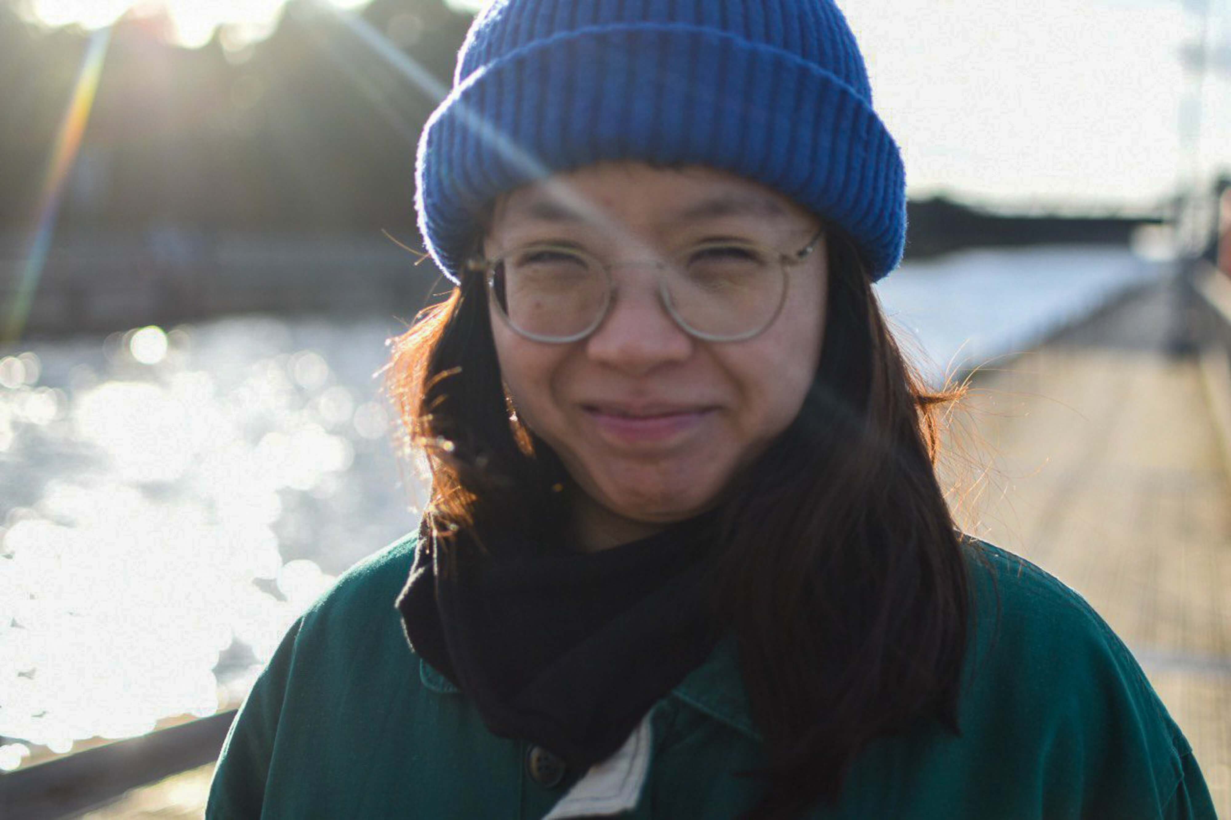 Porträt einer Frau mit Brille und blauer Strickmütze im Freien an einem Gewässer. Im Hintergrund sind Wasser, Uferpromenade und Sonnenlicht unscharf zu erkennen.