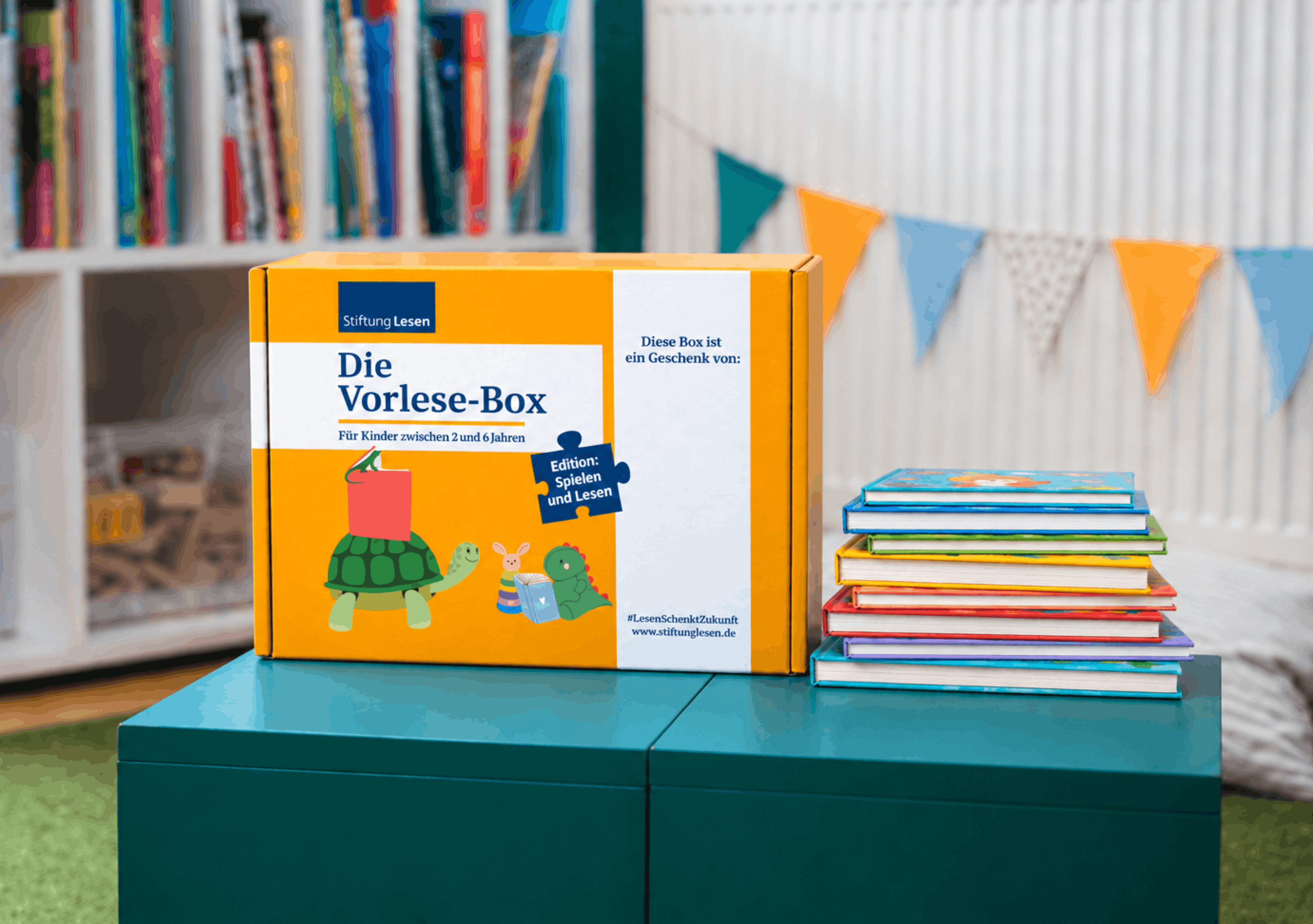 Orangefarbene Vorlese-Box der Stiftung Lesen für Kinder von zwei bis sechs Jahren, Edition „Spielen und Lesen“, neben einem Stapel bunter Kinderbücher. Im Hintergrund sind ein Bücherregal und eine Wimpelkette zu sehen.