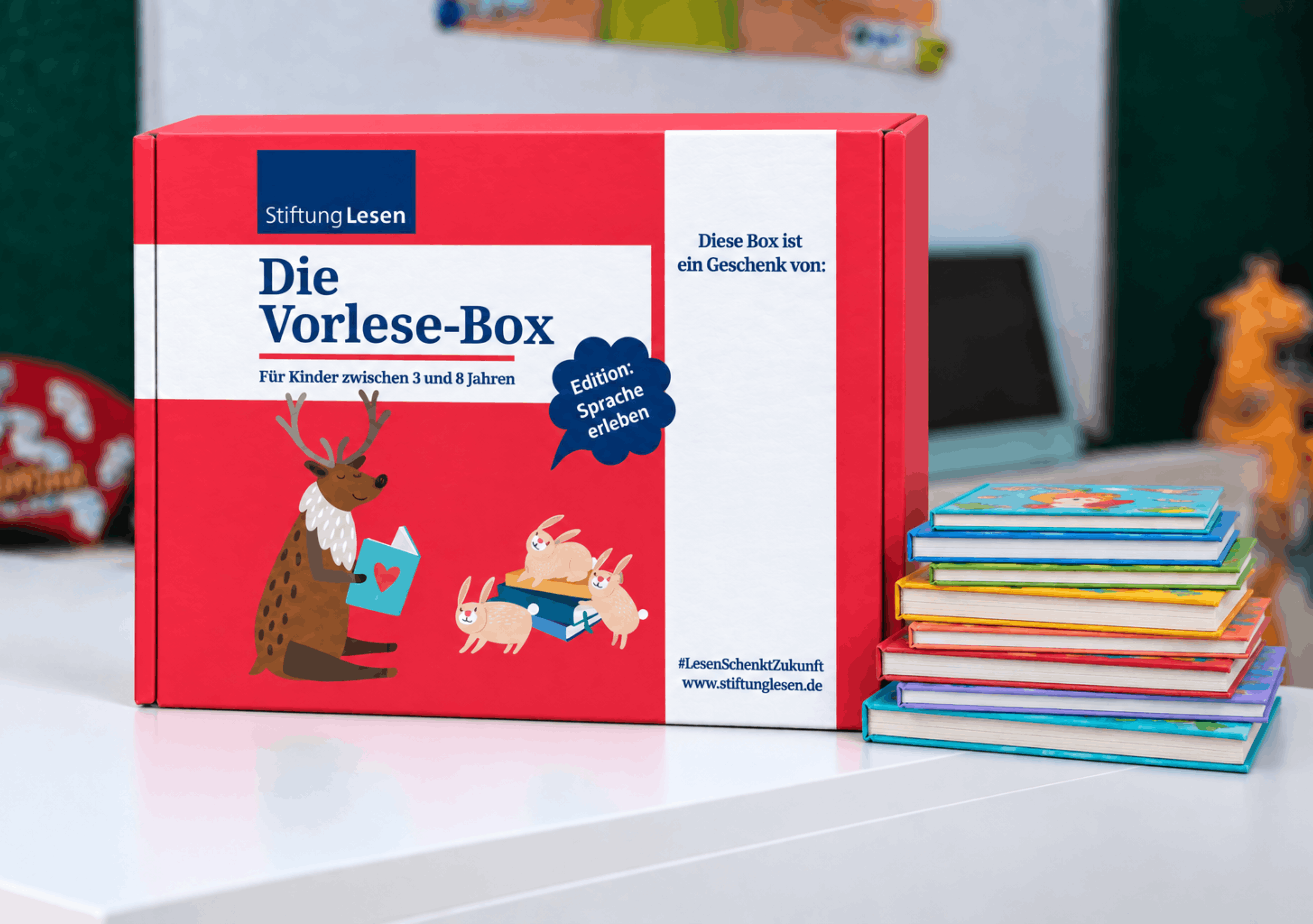 Rote Vorlese-Box der Stiftung Lesen für Kinder von drei bis acht Jahren, Edition „Sprache erleben“, neben einem Stapel bunter Kinderbücher. Die Box ist mit Illustrationen eines lesenden Rentiers und mehrerer Hasen gestaltet.