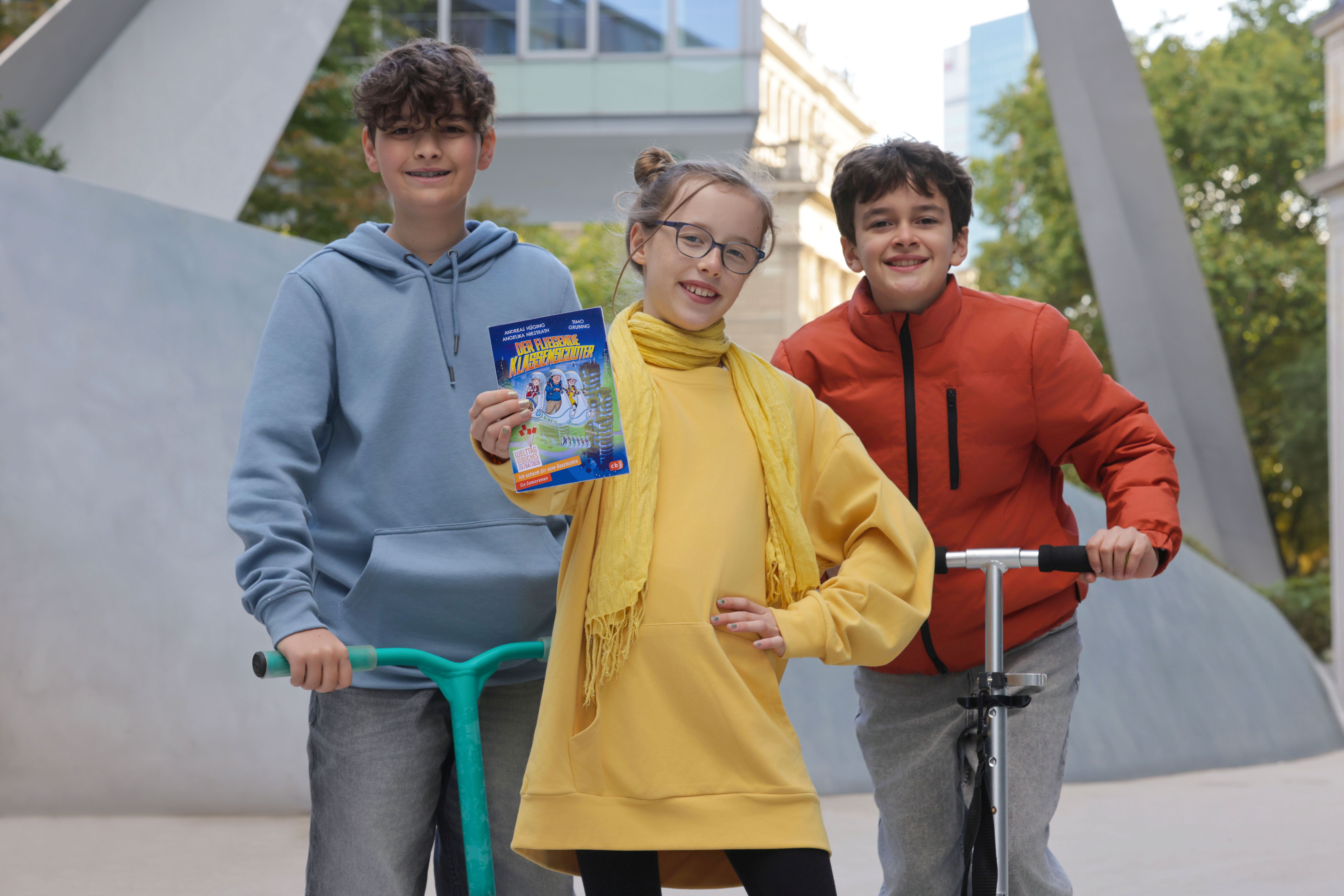 Drei Kinder stehen draußen in einer Stadtumgebung. Das Mädchen in der Mitte trägt ein gelbes Kleid und einen gelben Schal und hält ein Buch mit dem Titel „Klassenfahrt ins Abenteuer“ in die Kamera. Links steht ein Junge mit blauem Hoodie auf einem grünen Scooter, rechts ein Junge in roter Jacke mit einem silbernen Roller. Alle drei lächeln.