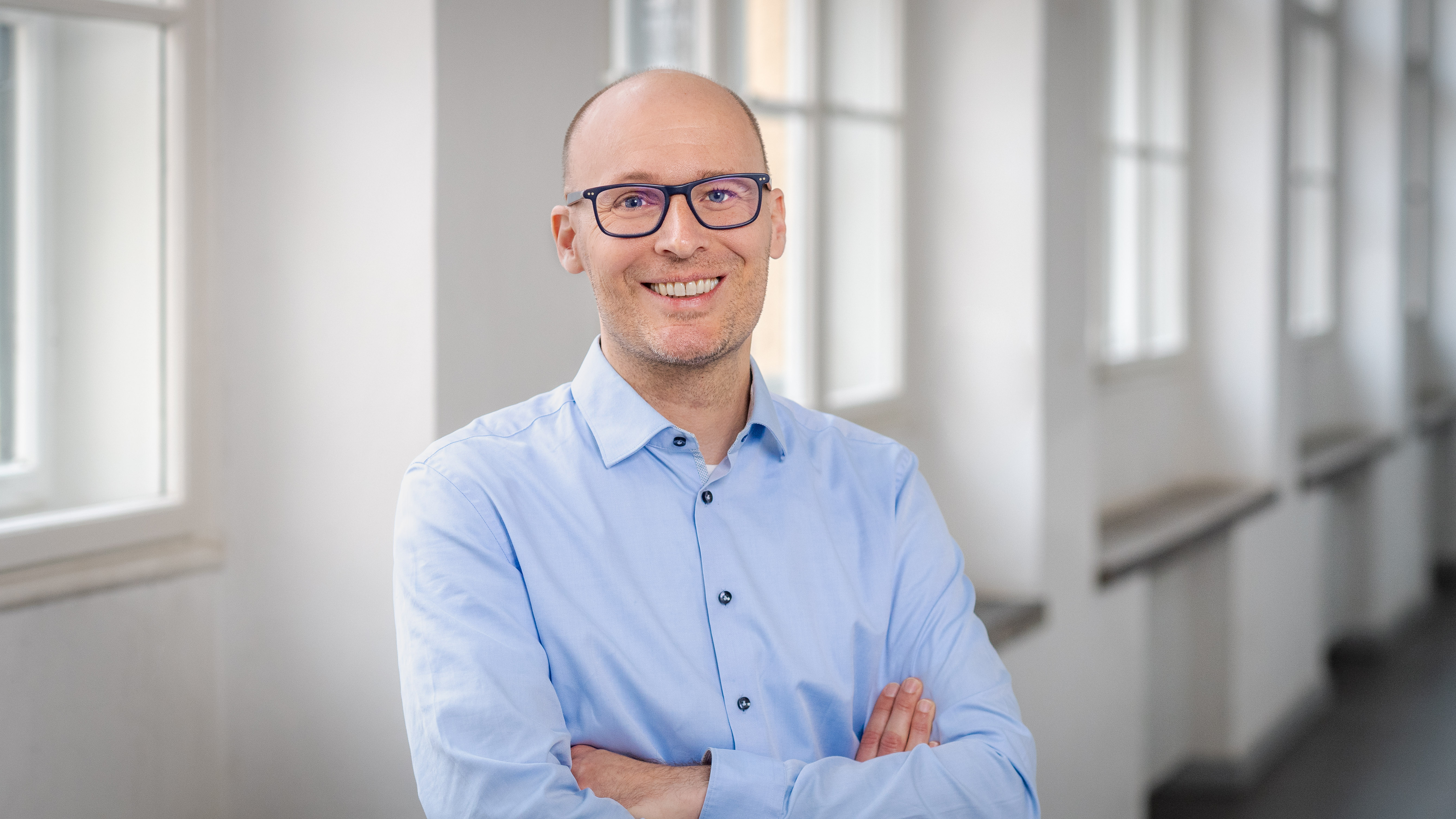 Porträt eines lächelnden Mannes mit Brille und hellblauem Hemd. Er steht mit verschränkten Armen in einem hellen Innenraum mit großen Fenstern; der Hintergrund ist unscharf.