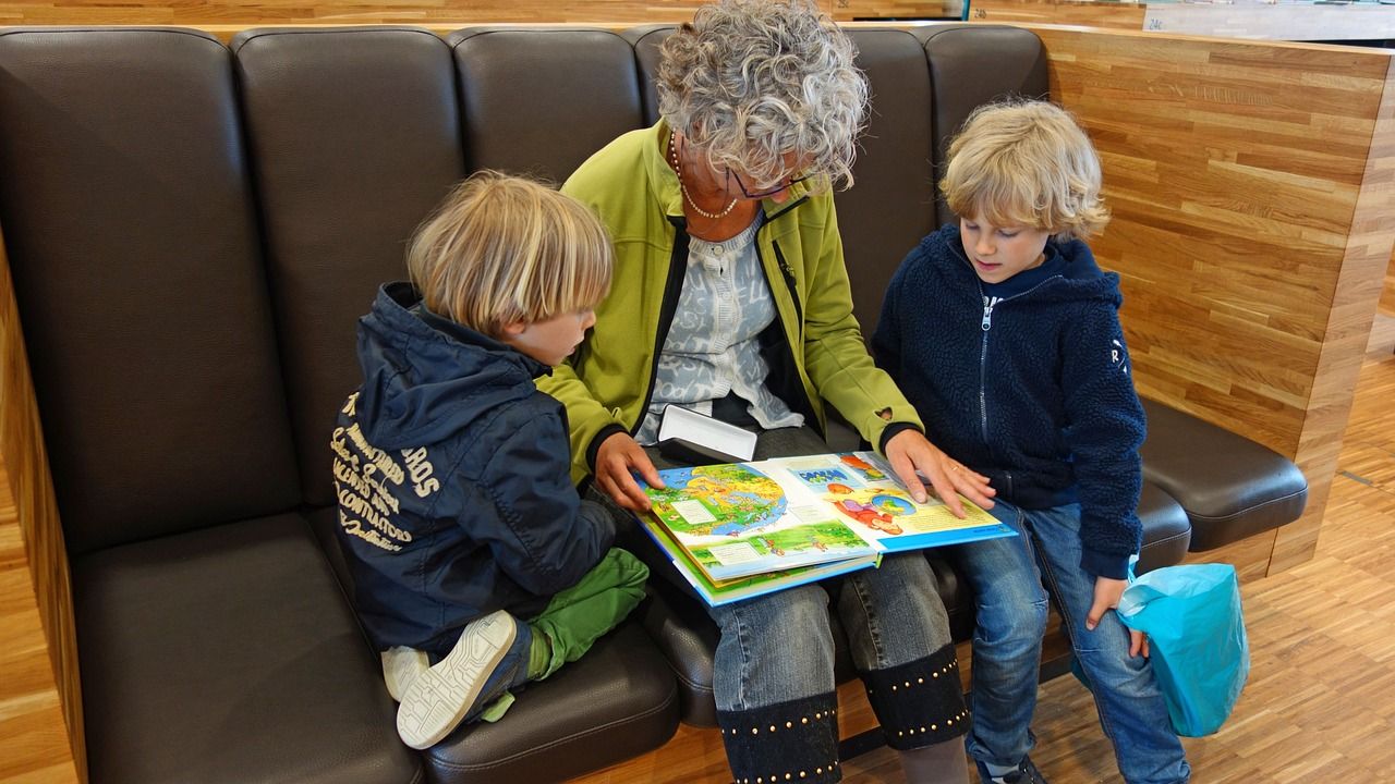 Eine älter Dame sitzt mit zwei Kindern auf einer gepolsterten Bank in einem öffentlichen Raum und liest gemeinsam mit den Kindern in einem Bilderbuch.