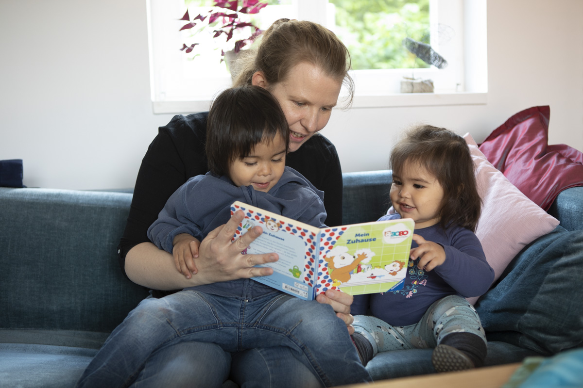 Eine Frau sitzt mit zwei Kindern im Alter von 2 und 3 Jahren auf der Couch und liest aus einem Buch vor