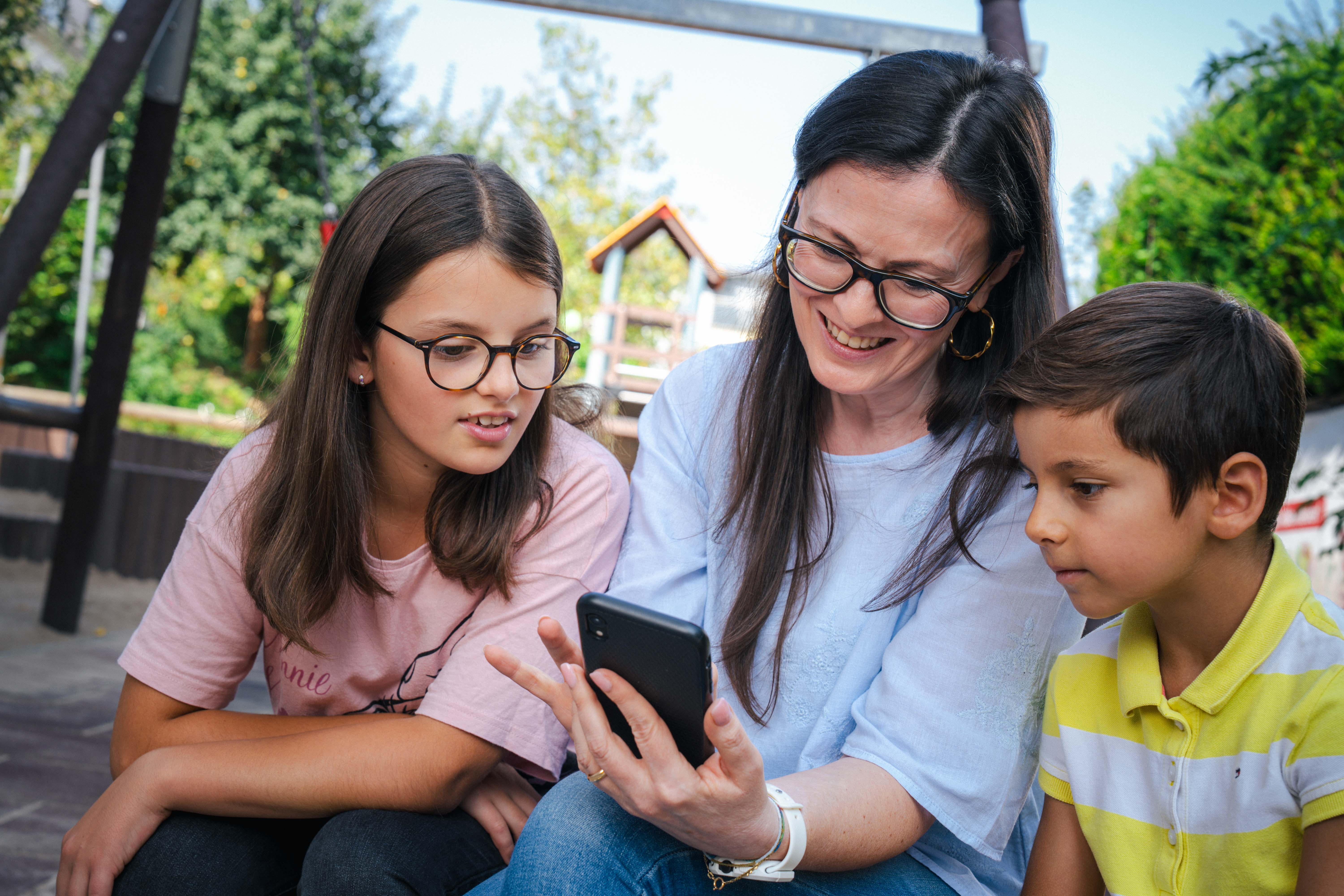 Ein Frau sitzt zwischen zwei Kindern auf einem Spielplatz. Sie hält ein Handy in der Hand, alle drei schauen auf das Display des Smartphones.