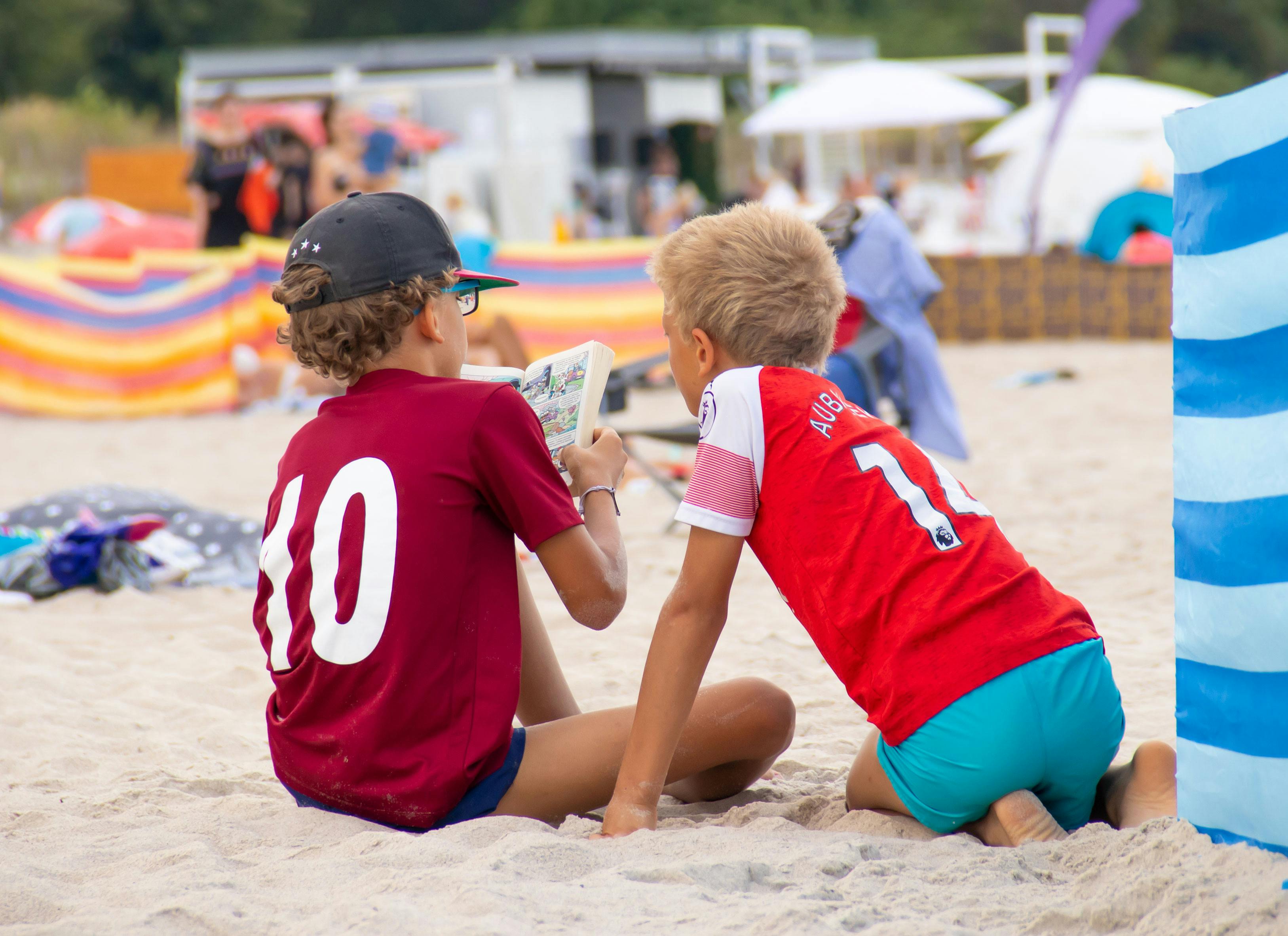 Zwei Jungs sitzen im Freibad im Sand und lesen in einem Comicheft.