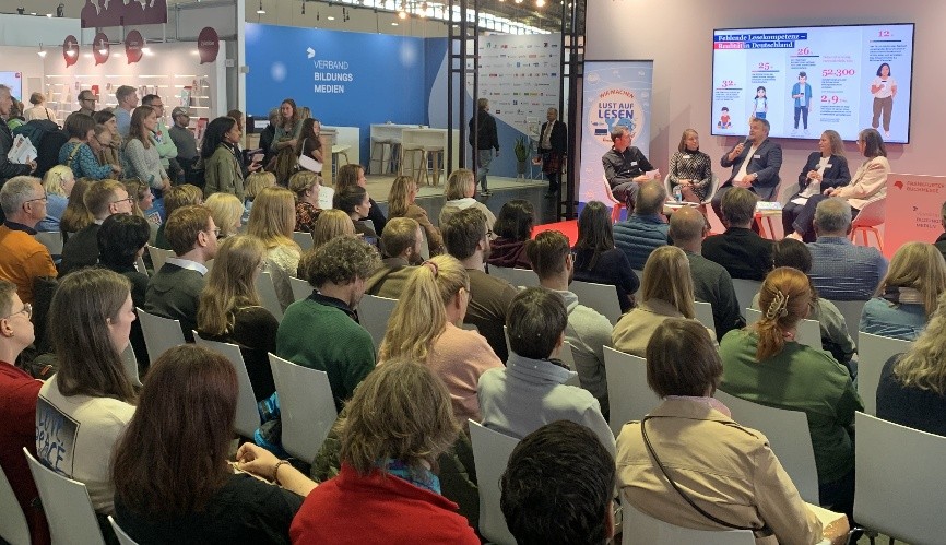 Foto einer Podiumsdiskussion mit vier Sprecherinnen vor Publikum auf einer Messe oder Veranstaltung. Im Hintergrund ist ein Bildschirm mit Infografik und ein Banner mit der Aufschrift „Lust am Lesen“ zu sehen. Viele Zuhörerinnen verfolgen aufmerksam das Gespräch