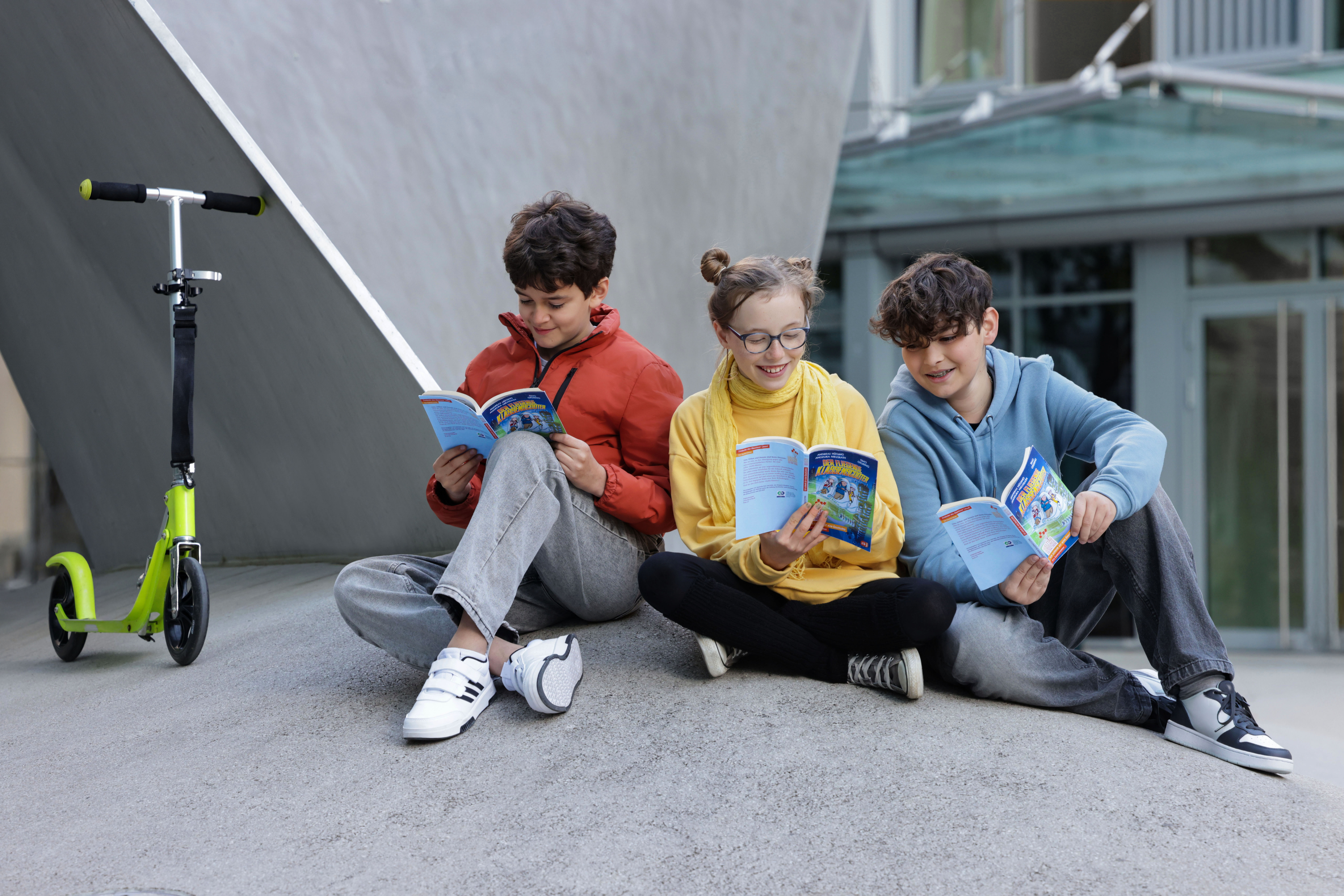 Drei Kinder sitzen neben einem Scooter auf einer futuristisch anmutenden Betonlandschaft und lesen im aktuellen Welttagsbuch