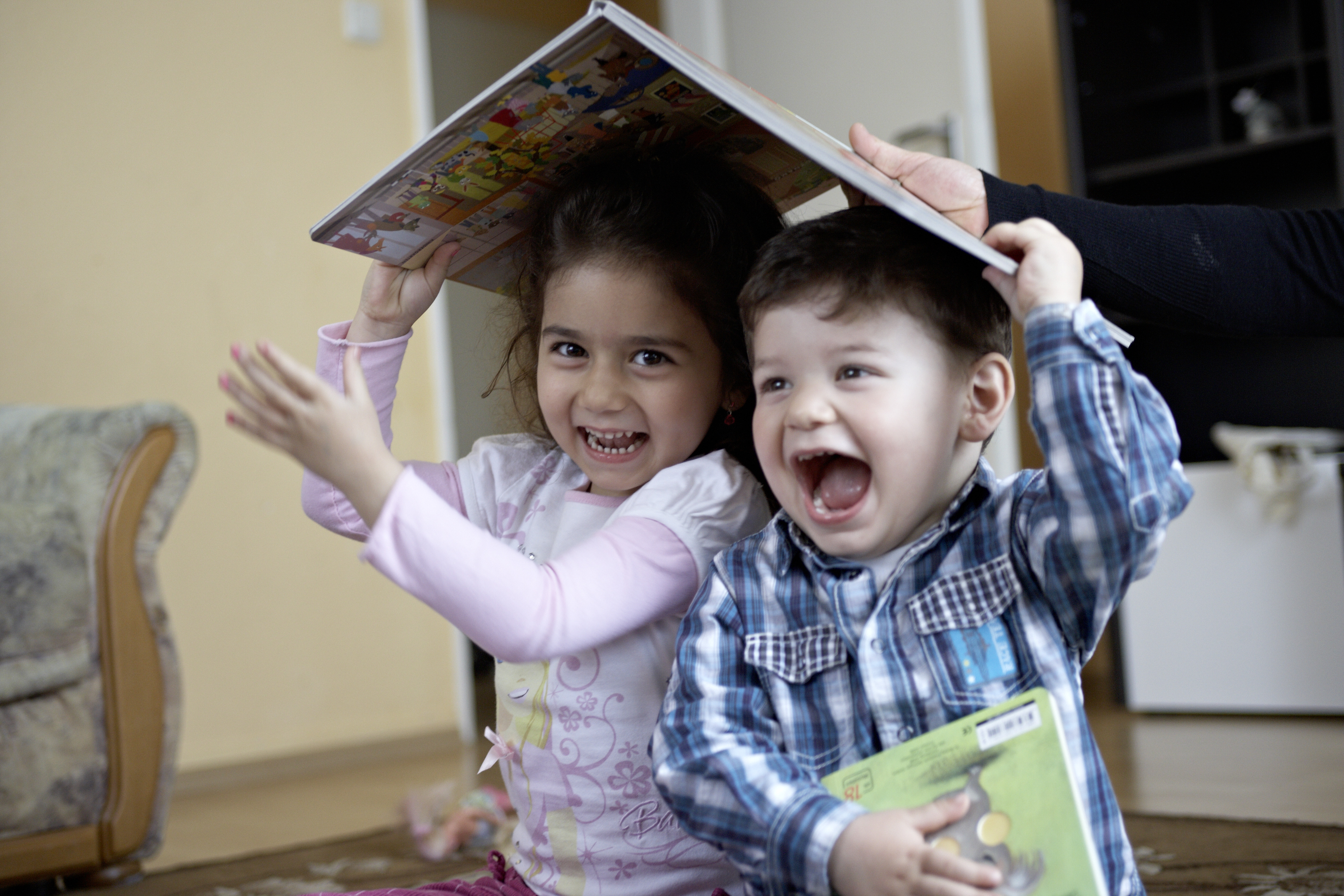 Zwei Kleinkinder sitzen lachend auf dem Boden und halten sich ein Buch wie ein Dach über den Kopf