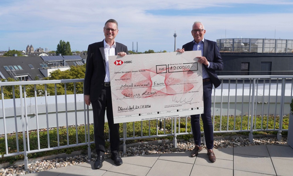 Zwei Männer stehen auf einer Dachterrasse und halten gemeinsam einen großen Spendenscheck von HSBC in die Kamera. Im Hintergrund sind Gebäude und blauer Himmel zu sehen.