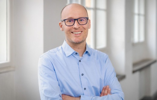 Porträt eines lächelnden Mannes mit Brille und hellblauem Hemd. Er steht mit verschränkten Armen in einem hellen Innenraum mit großen Fenstern.