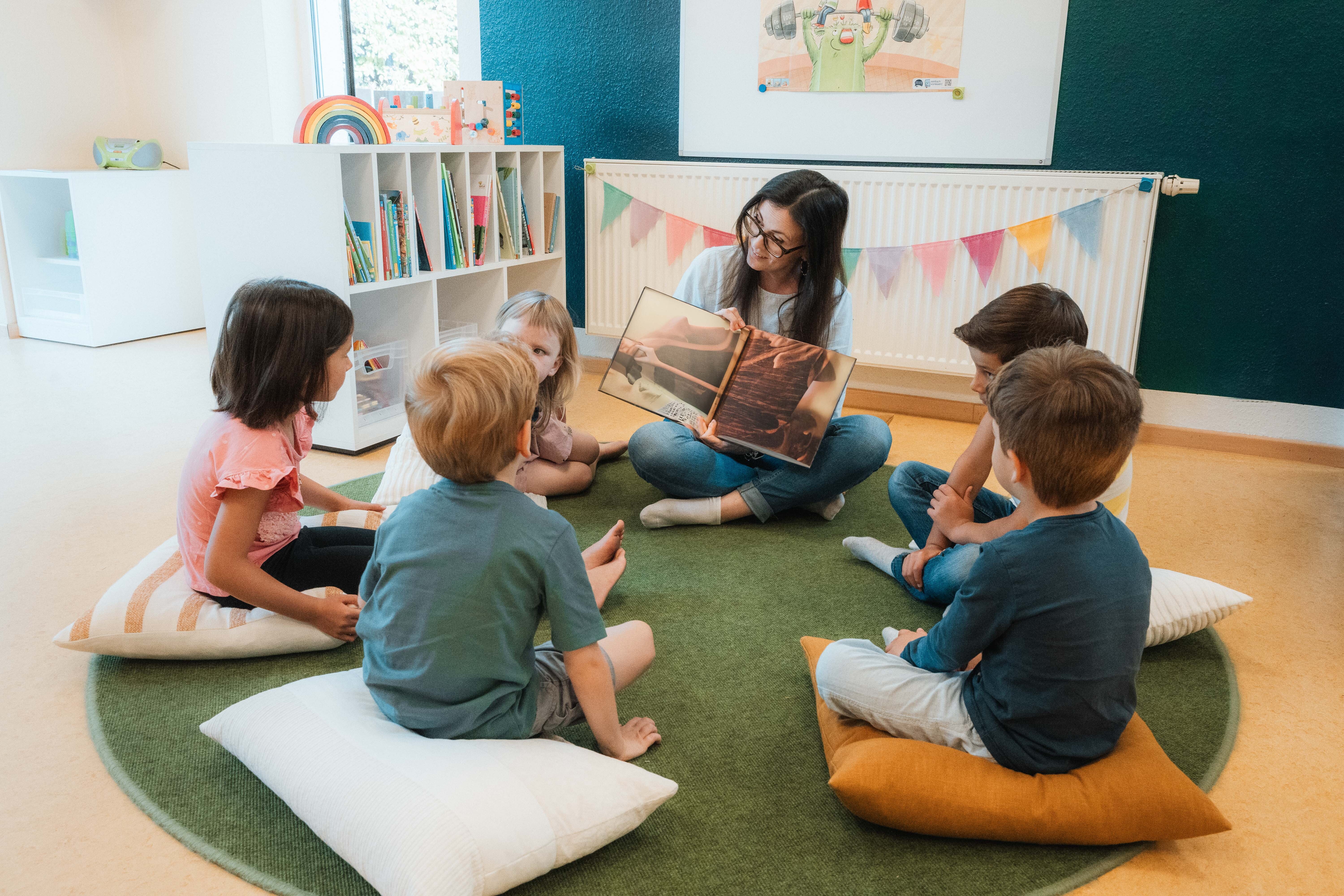 Eine Frau liest einer Gruppe von fünf Kindern aus einem Buch vor. Die Frau und die Kinder sitzen in einer Kita in einem Kreis. Die Kinder hören aufmerksam zu.