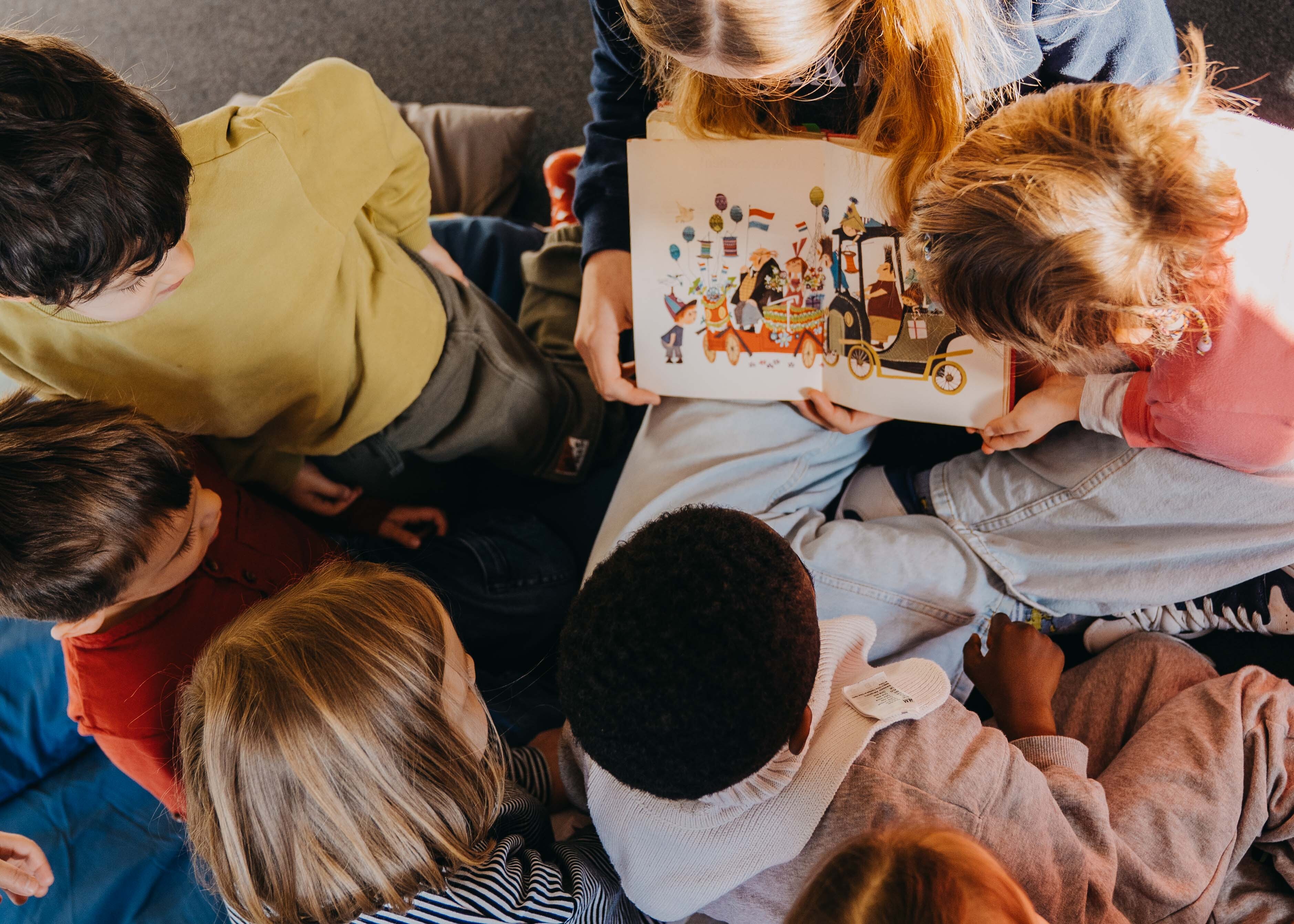 Eine Gruppe von Kindern sitzt im Kreis um eine erwachsene Person, die ein bunt illustriertes Bilderbuch aufgeschlagen hält. Die Aufnahme aus der Vogelperspektive zeigt, wie die Kinder gemeinsam die Bilder betrachten und einer Vorlesegeschichte folgen.  