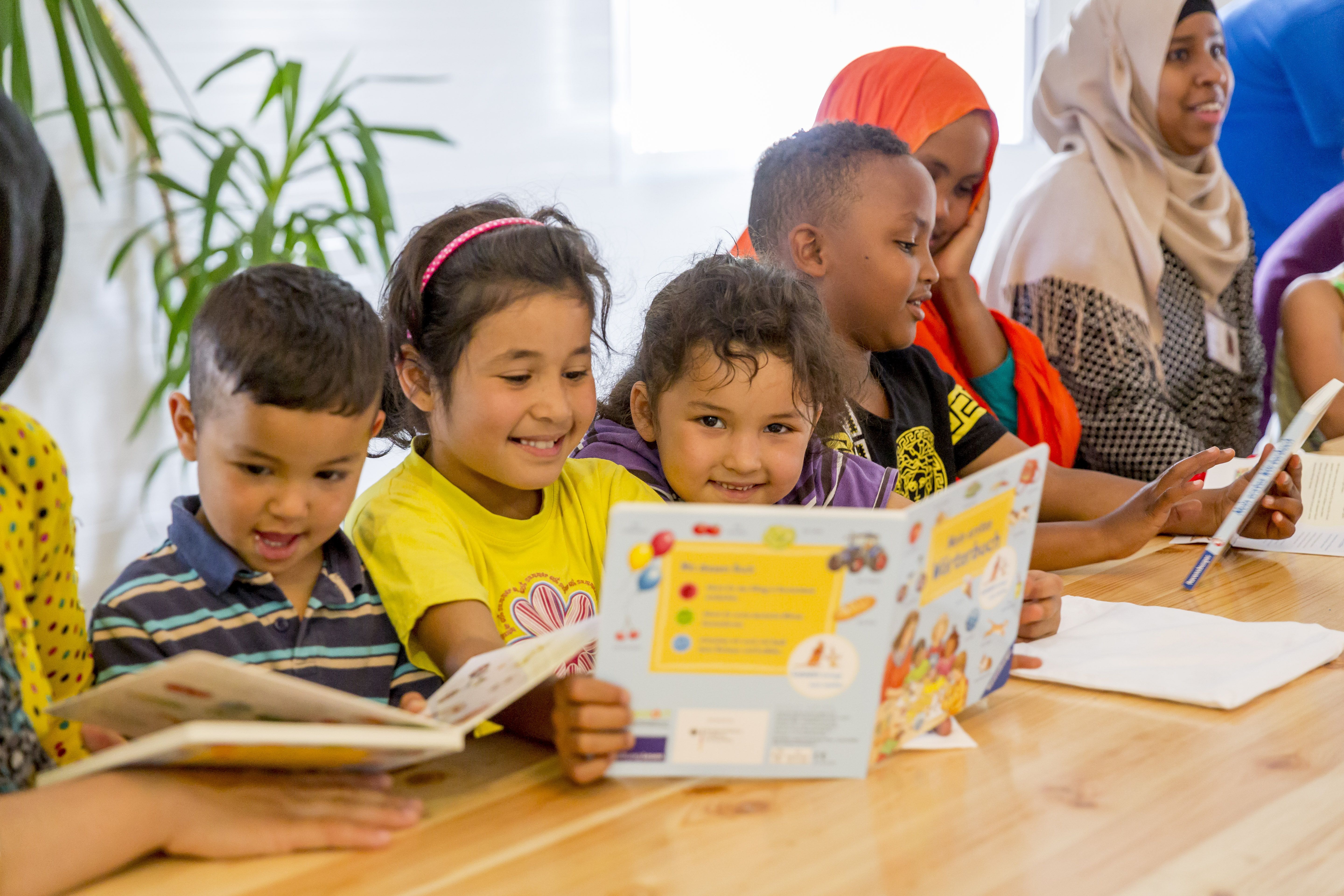 Kindergruppe liest gemeinsam Bücher an einem Holztisch in einem hellen Raum.