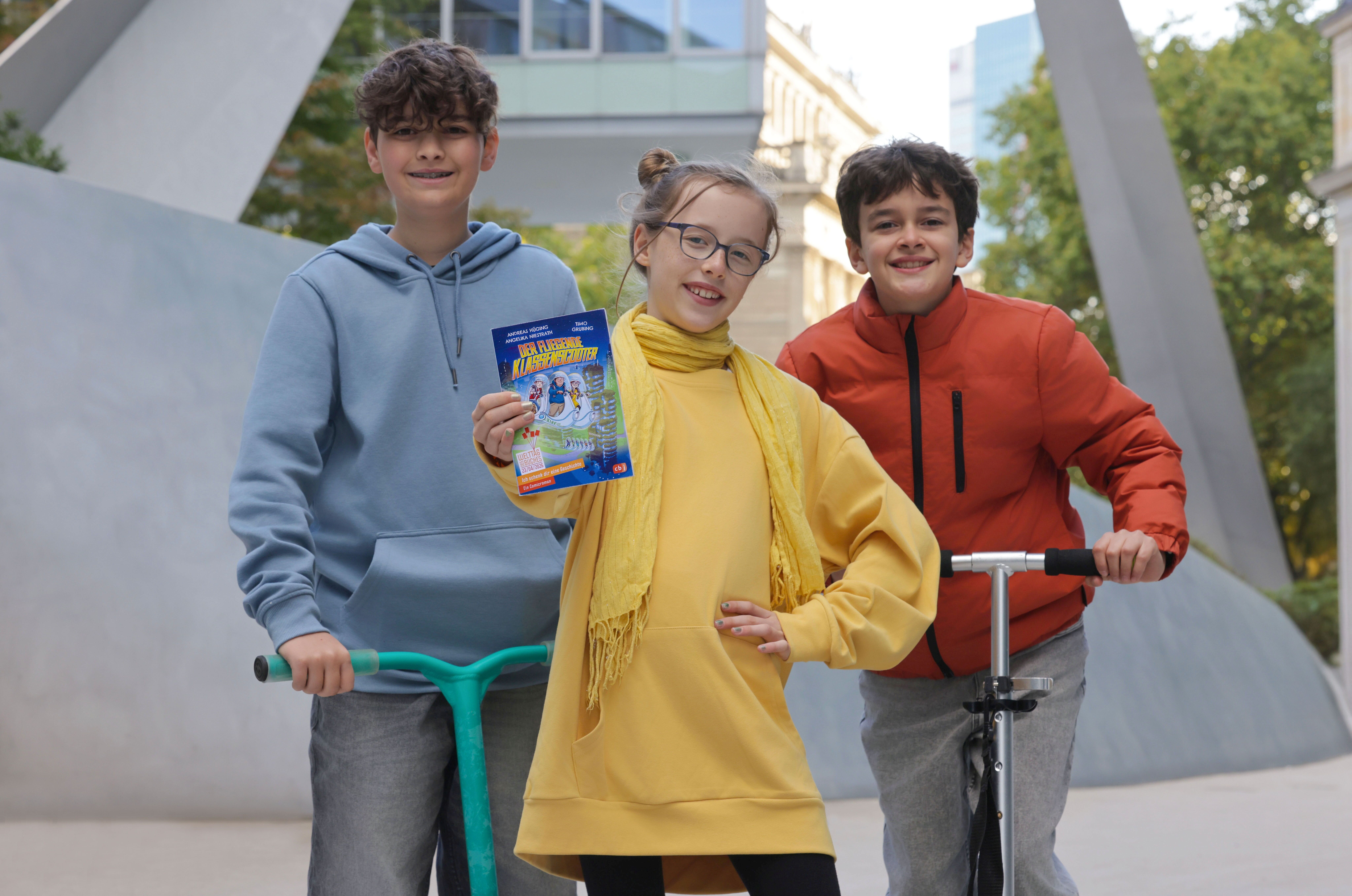 Drei Kinder stehen draußen in einer Stadtumgebung. Das Mädchen in der Mitte trägt ein gelbes Kleid und einen gelben Schal und hält ein Buch mit dem Titel „Klassenfahrt ins Abenteuer“ in die Kamera. Links steht ein Junge mit blauem Hoodie auf einem grünen Scooter, rechts ein Junge in roter Jacke mit einem silbernen Roller. Alle drei lächeln.