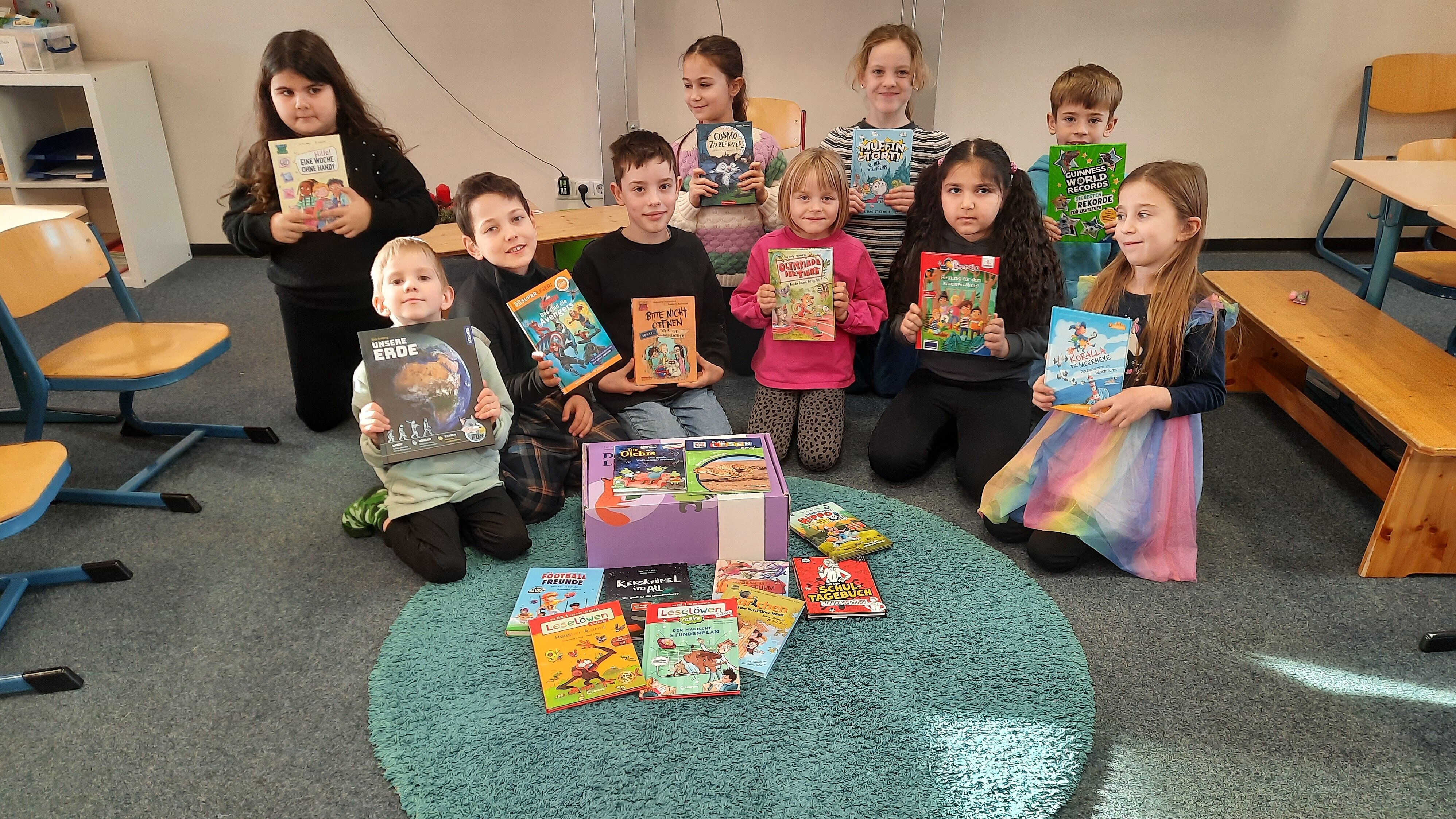 Zehn Grundschulkinder sitzen auf dem Boden und lächeln in die Kamera. Sie halten verschiedene Kinderbücher in den Händen, die sie fröhlich präsentieren. Vor ihnen liegen eine lila "Lese-Box" und einige Bücher auf dem Boden.