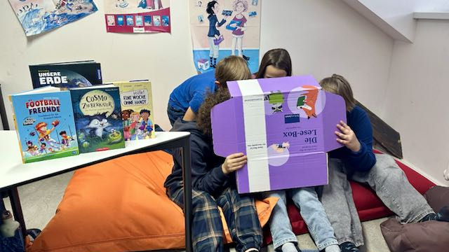 Mehrere Kinder sitzen dicht beieinander auf Kissen und lesen gemeinsam aus einer großen lila Lesebox. Daneben steht ein Tisch mit verschiedenen Kinderbüchern, im Hintergrund sind Poster und Zeichnungen an der Wand zu sehen.