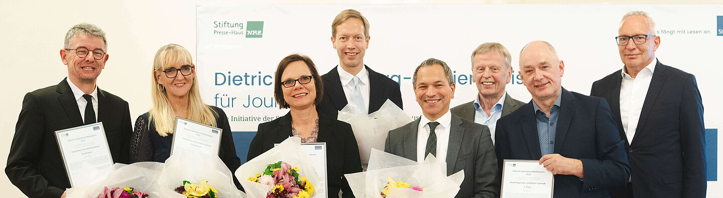 Gruppenfoto von acht Personen bei der Verleihung des Dietrich-Oppenberg-Medienpreises 2026. Die Preisträgerinnen und Preisträger halten Blumensträuße und Urkunden in den Händen. Im Hintergrund ist eine Veranstaltungswand mit dem Schriftzug des Medienpreises zu sehen.