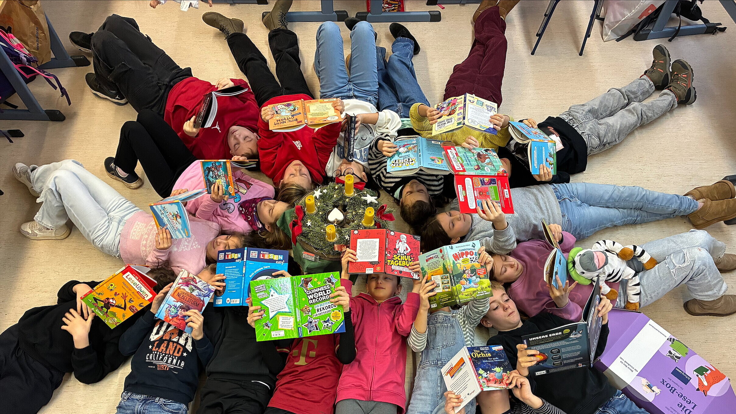 Mehrere Grundschulkinder liegen im Kreis auf dem Boden eines Klassenzimmers und lesen Kinderbücher. In der Mitte liegt ein Adventskranz, rundherum halten die Kinder ihre Bücher über sich oder neben sich, einige schauen lachend in die Bücher. Die Szene vermittelt gemeinsames Lesen in entspannter Atmosphäre.