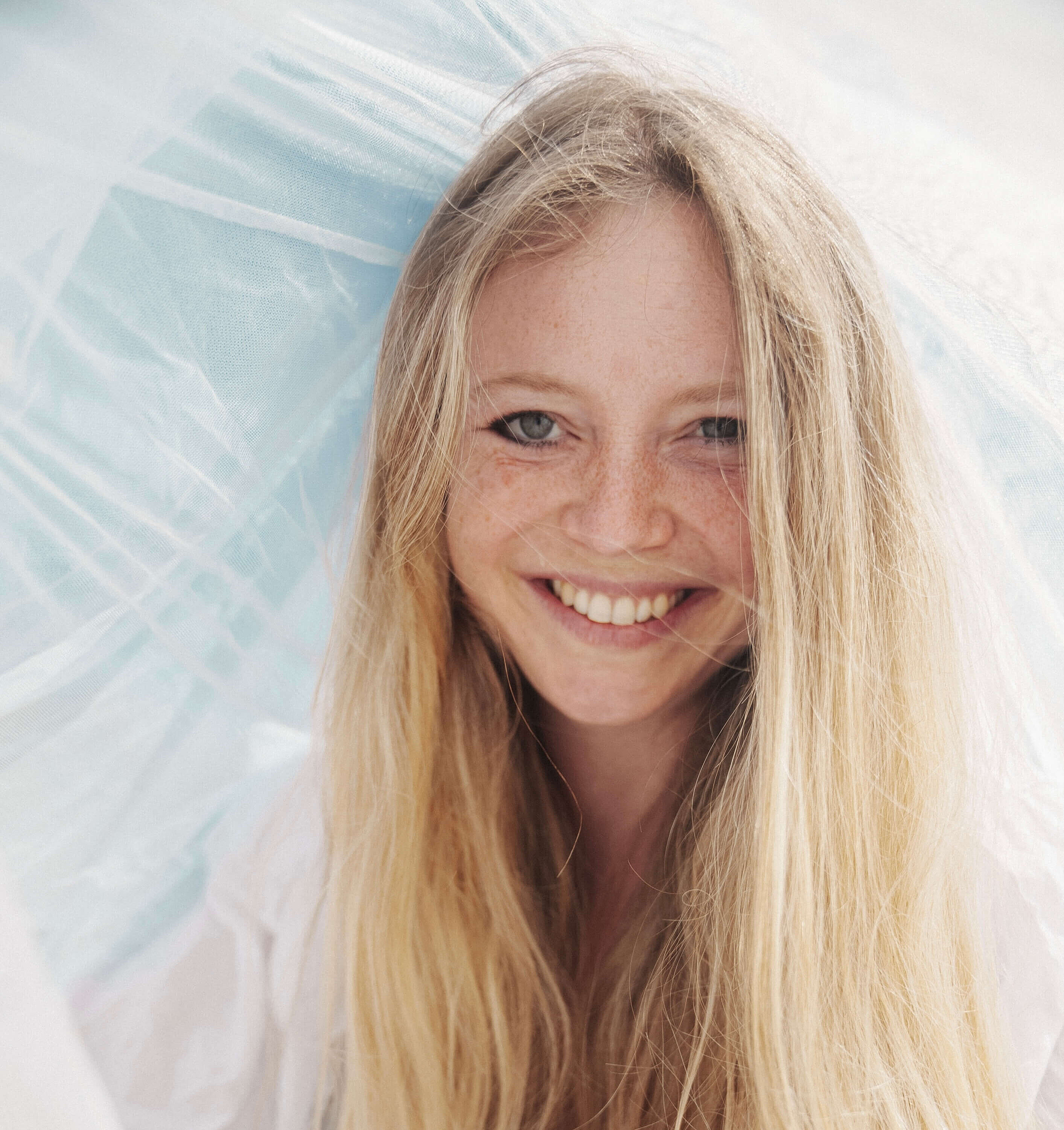 Porträt eines lächelnden Mädchens mit langen blonden Haaren und Sommersprossen, vor hellem, weich strukturiertem Hintergrund.