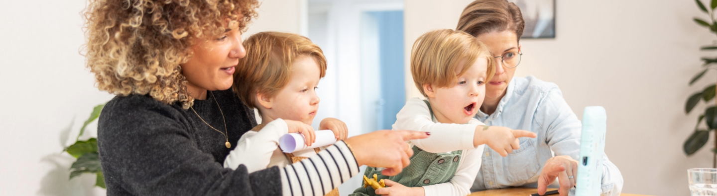 Mehrere erwachsene Personen sitzen mit zwei Kindern am Tisch und schauen gemeinsam auf einen Gegenstand vor ihnen, während sie in dieselbe Richtung zeigen.“