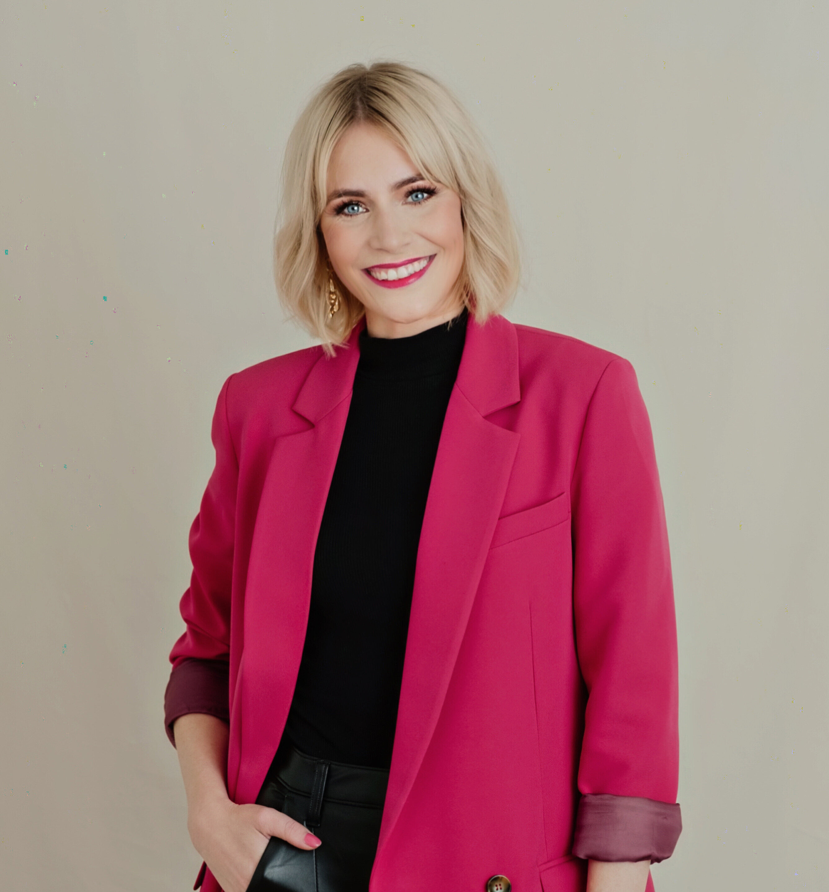 Porträt einer lächelnden Frau mit blondem Haar, die einen pinken Blazer und schwarze Kleidung trägt und in die Kamera blickt.