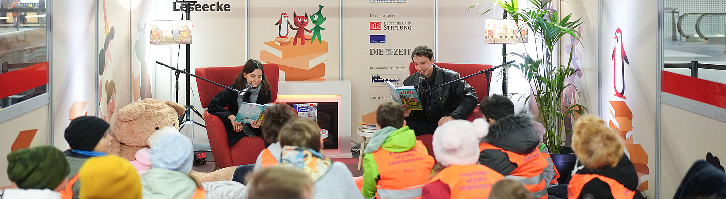 Zwei Personen lesen Kindern in einer eingerichteten Leseecke im Bahnhof vor; die Kinder sitzen in Gruppen davor und tragen orangefarbene Westen.