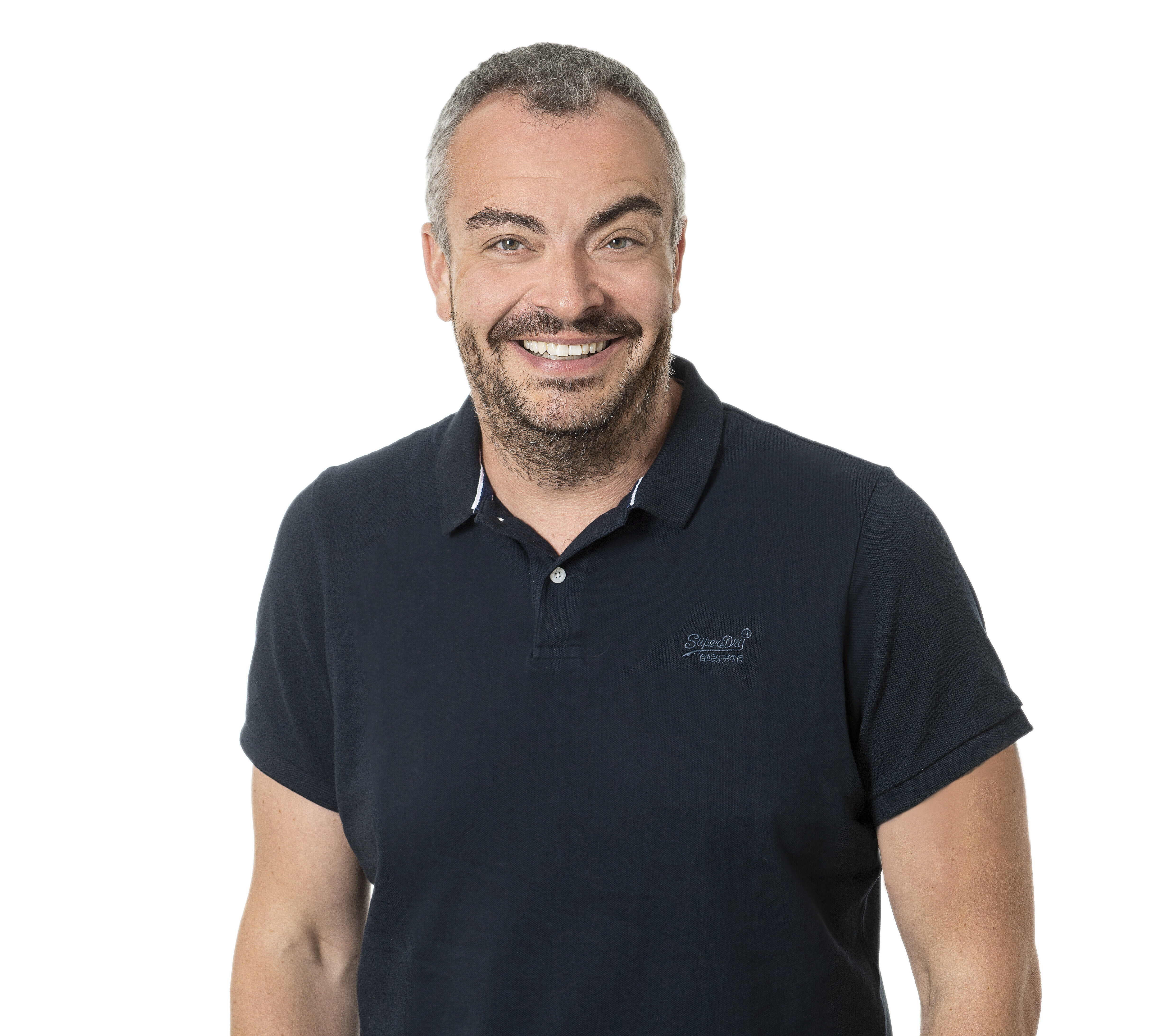 Eine stehende Person trägt ein dunkles, kurzärmliges Poloshirt und eine Armbanduhr mit farbigem Armband. Die Person hat die Hände in den Hosentaschen und steht vor weißem Hintergrund.