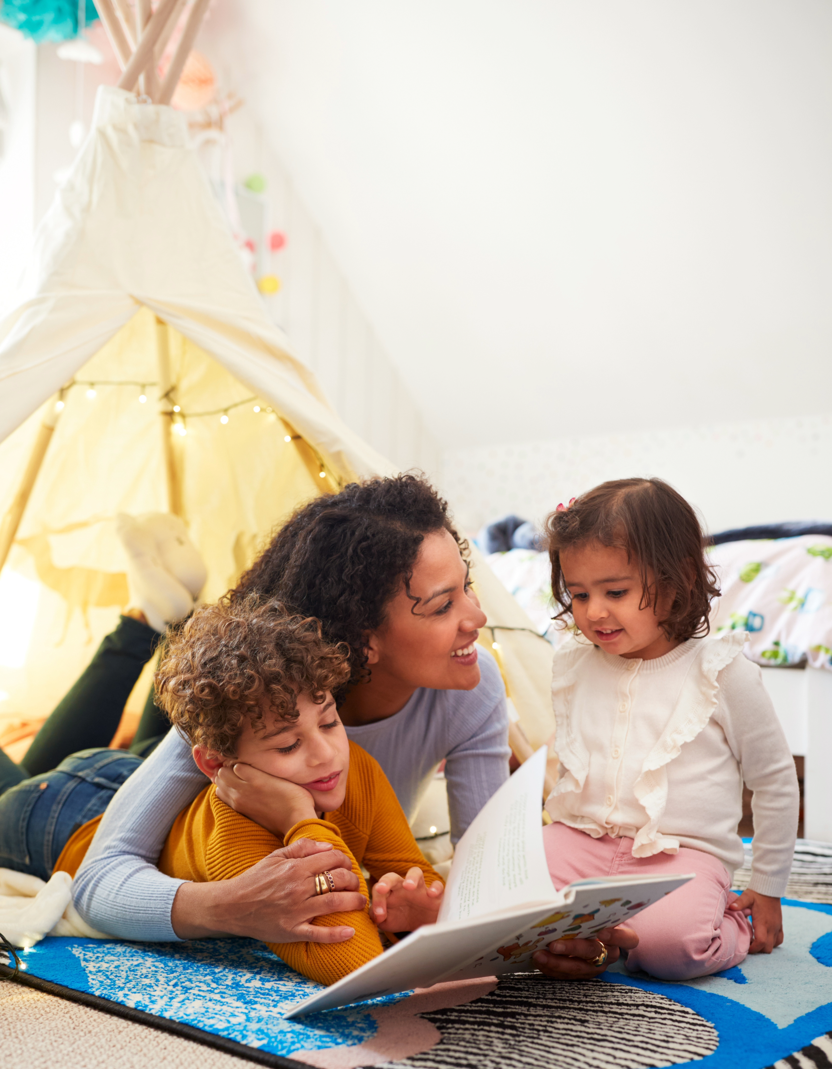 Eine Erwachsene liest zwei Kindern auf einem Teppich aus einem Bilderbuch vor. Die drei schauen gemeinsam auf die geöffneten Seiten. Im Hintergrund steht ein beleuchtetes Spielzelt, das eine gemütliche Vorleseatmosphäre schafft.
