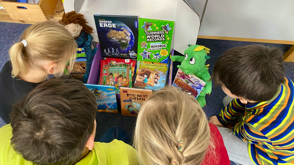 Mehrere Kinder stehen um eine geöffnete Lese-Box und schauen gemeinsam auf die darin liegenden Kinderbücher; ein Kuscheltier liegt zwischen den Büchern auf dem Boden.
