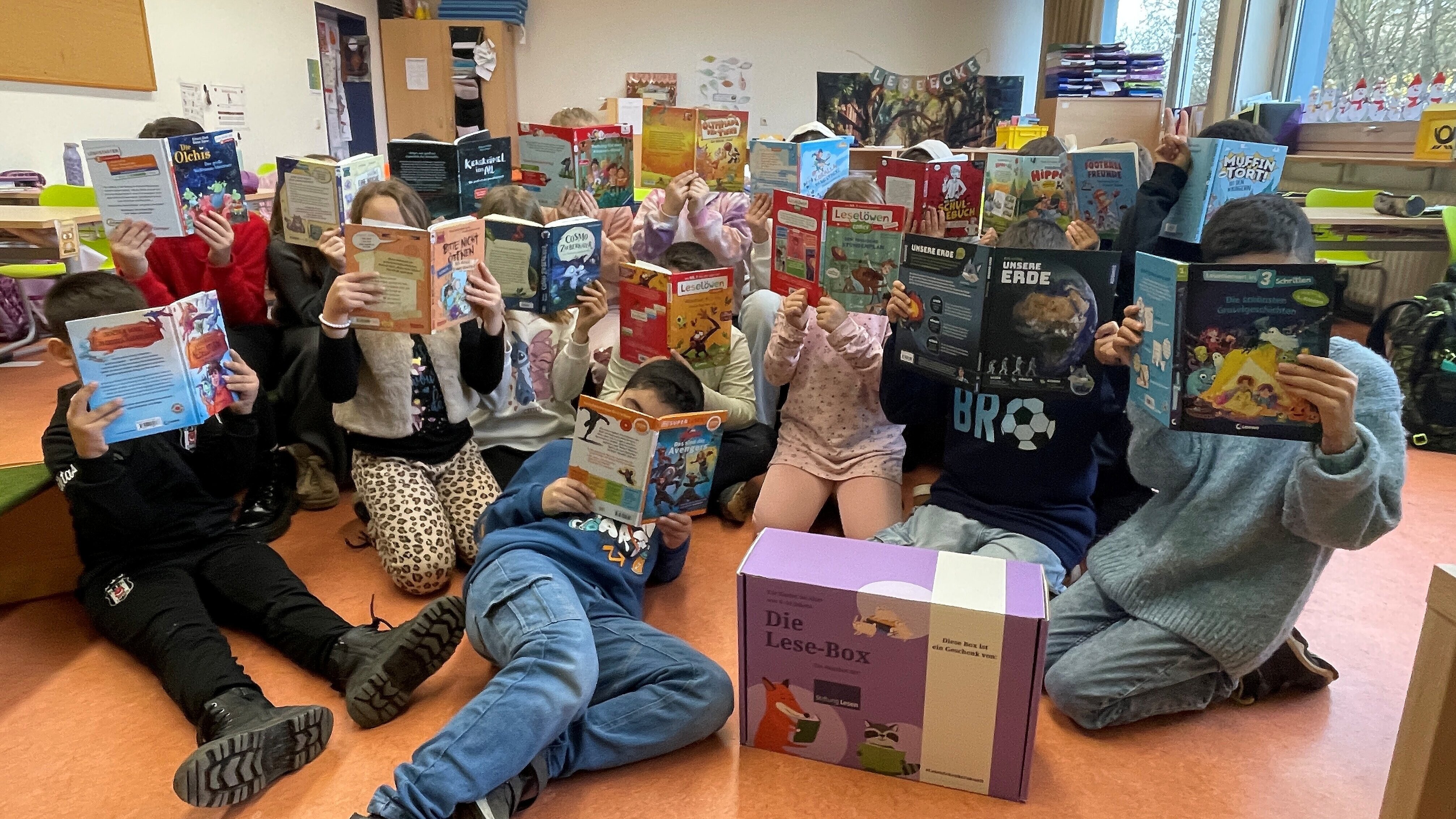 Eine große Gruppe von Kindern sitzt und kniet in einem Klassenraum auf dem Boden und hält aufgeklappte Kinderbücher vor ihr Gesicht. Vor ihnen steht eine lila „Lese-Box“. Im Hintergrund sind Tische, Regale und Unterrichtsmaterialien zu sehen.