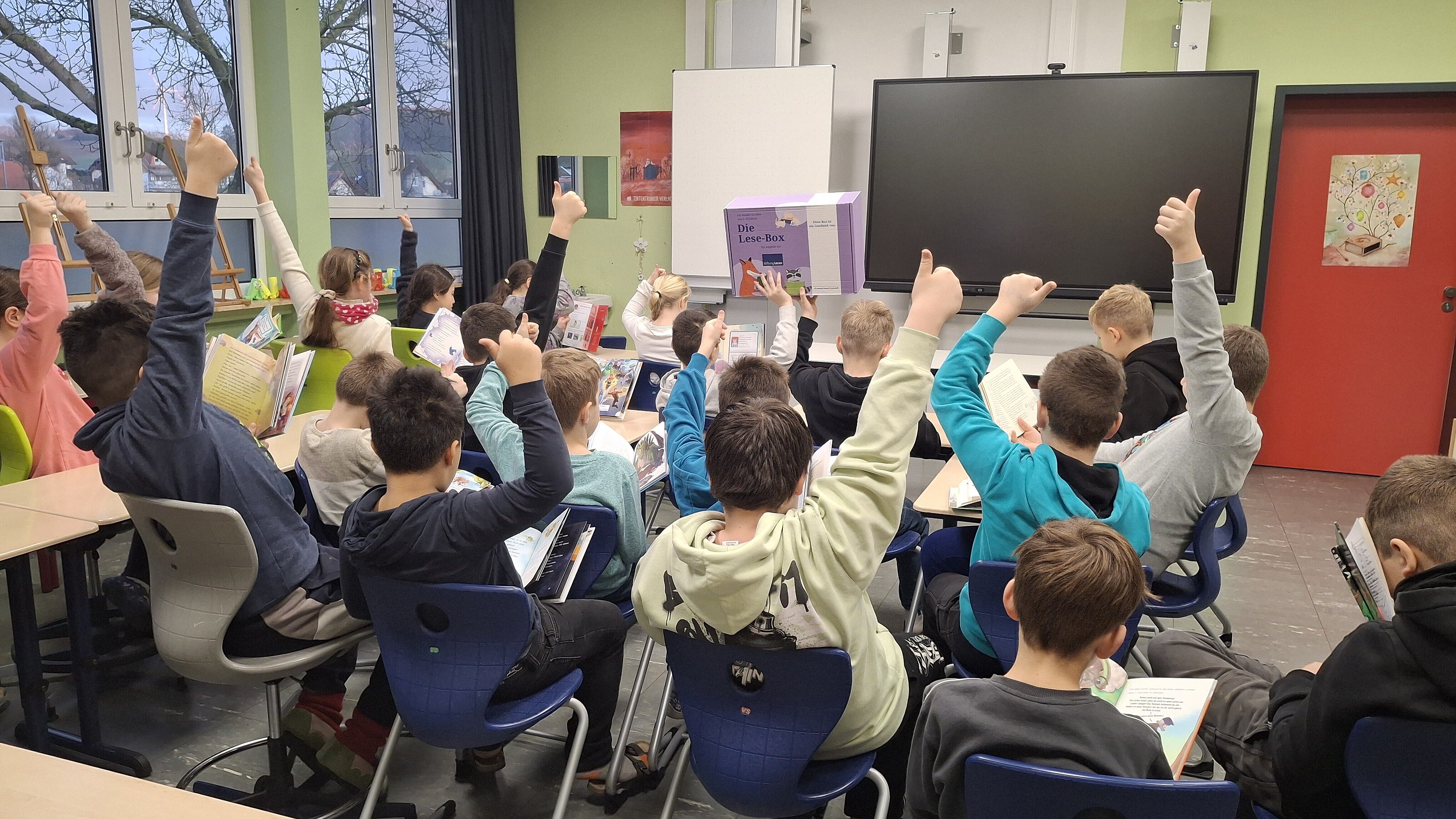 Grundschulklasse im Klassenzimmer: Kinder sitzen an Tischen, lesen in Büchern und strecken begeistert den Daumen nach oben. Im Hintergrund steht eine „Die Lese-Box“ vor einer digitalen Tafel.
