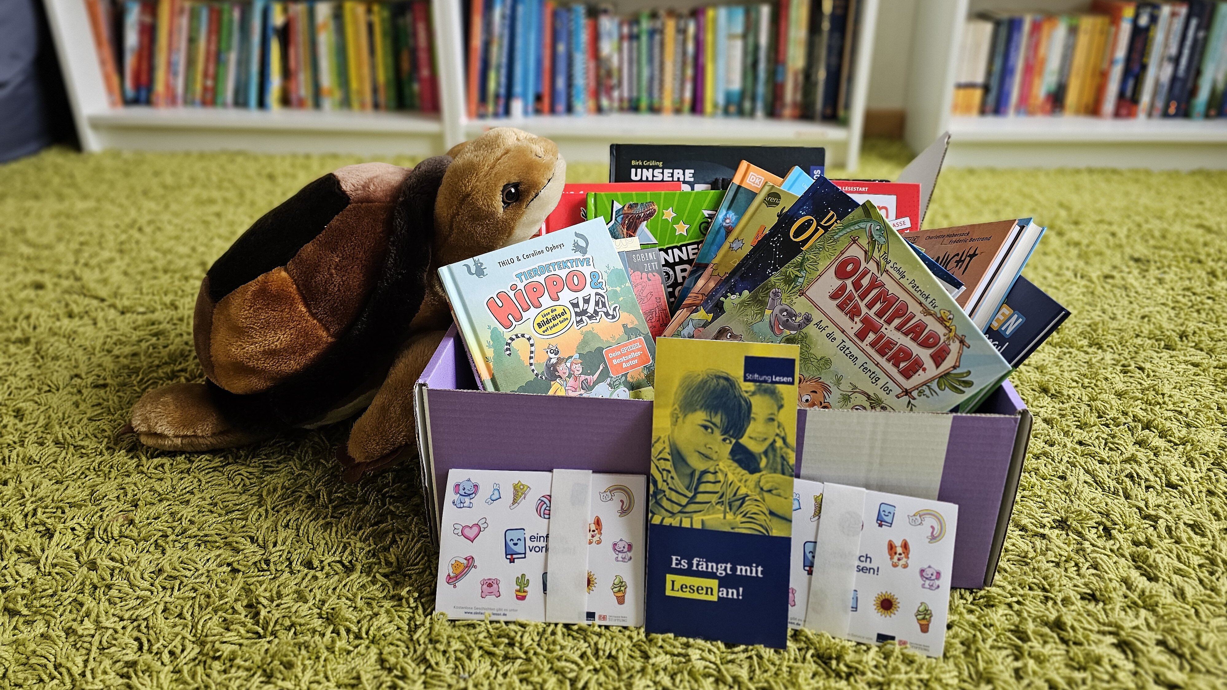 Eine geöffnete „Lese-Box“ mit verschiedenen Kinderbüchern steht auf einem grünen Teppich in einer Bibliothek; daneben liegt ein braunes Stofftier, im Hintergrund sind Bücherregale mit Kinder- und Jugendbüchern zu sehen.