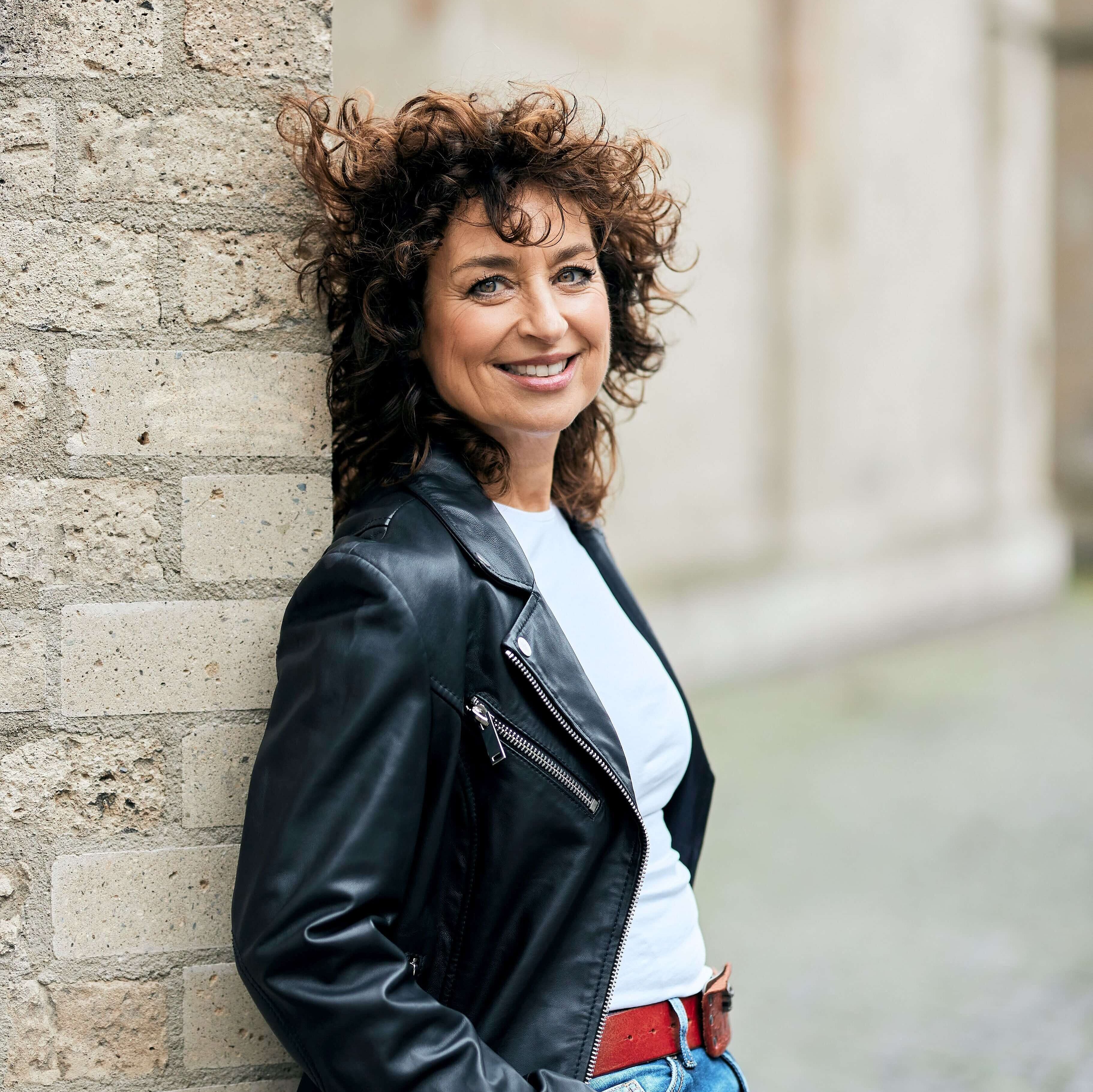 Porträt von Isabel Varell in Lederjacke, die an einer hellen Steinwand lehnt.