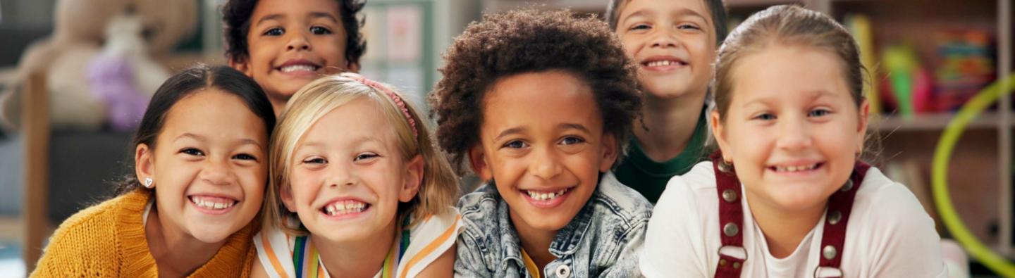 Sechs Kinder unterschiedlichen Aussehens liegen nebeneinander auf dem Bauch und lächeln fröhlich in die Kamera. Sie sitzen in einem gemütlichen Raum, im Hintergrund sind Spielzeug und Regale zu sehen.