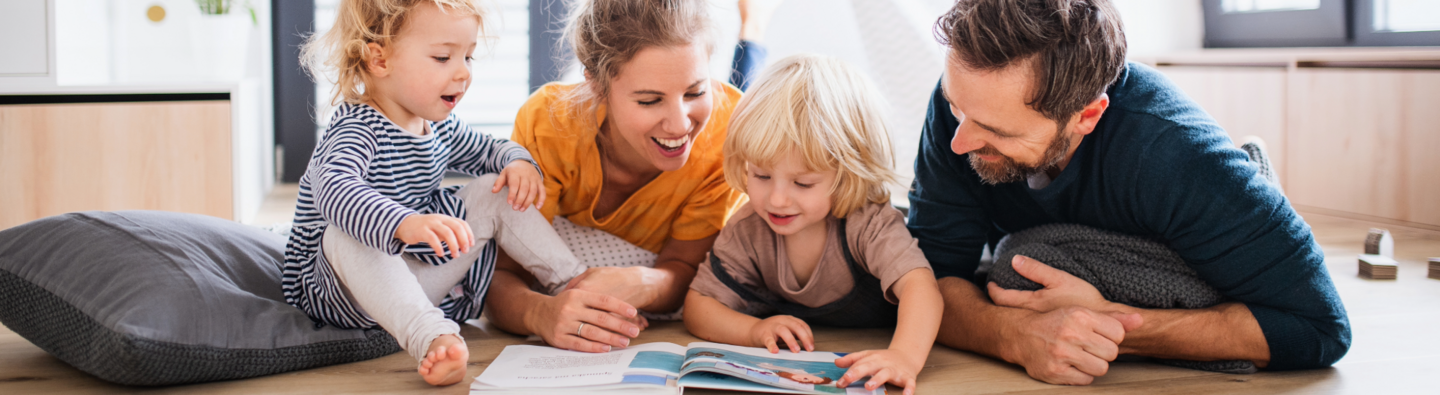 Familie liegt gemeinsam auf dem Boden und schaut mit zwei kleinen Kindern in ein Bilderbuch.
