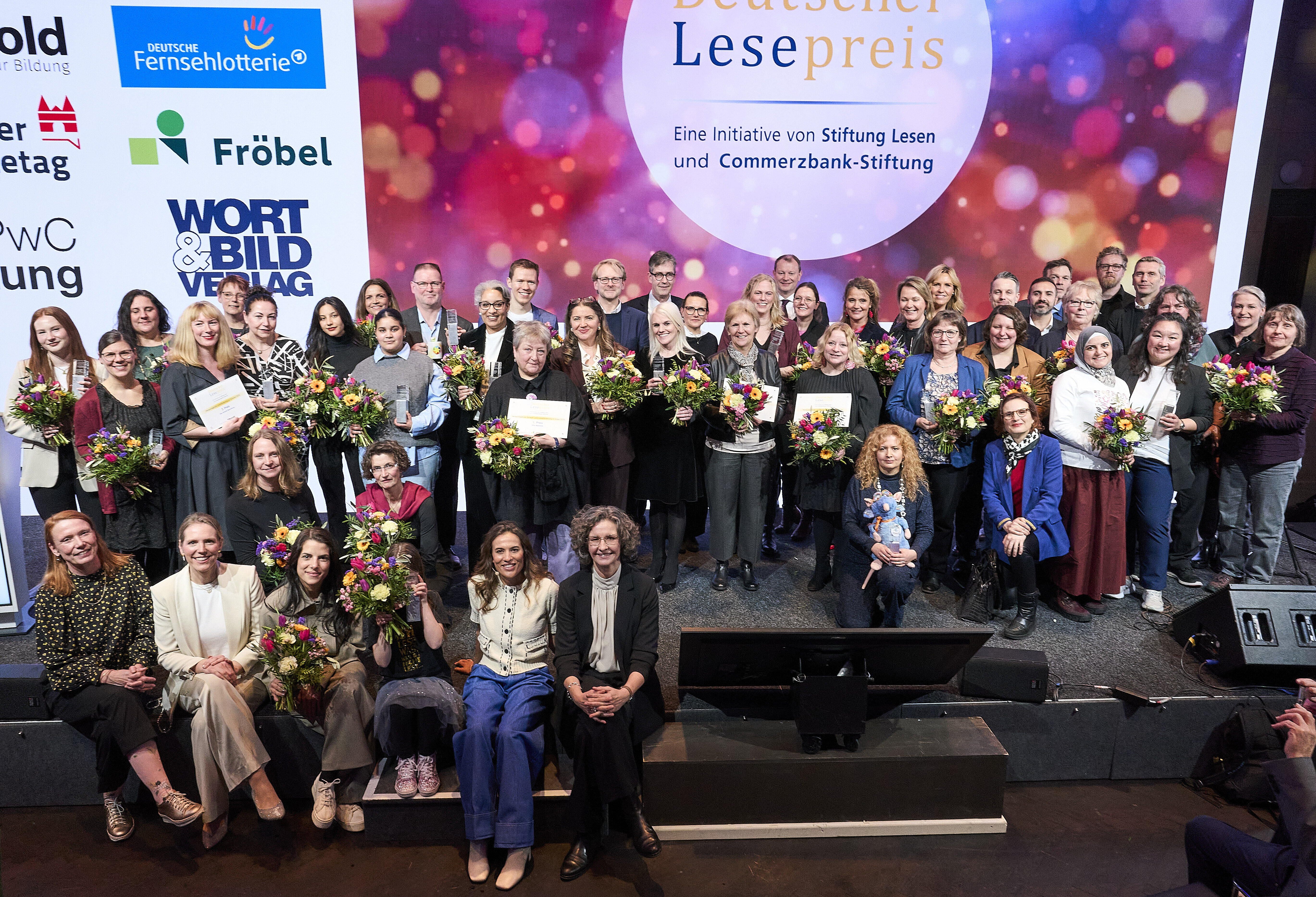 Große Gruppe von Preisträger*innen steht gemeinsam auf einer Bühne beim Deutschen Lesepreis, viele halten Blumensträuße und Urkunden in den Händen; im Hintergrund ist eine Veranstaltungswand mit dem Schriftzug „Deutscher Lesepreis“ zu sehen.