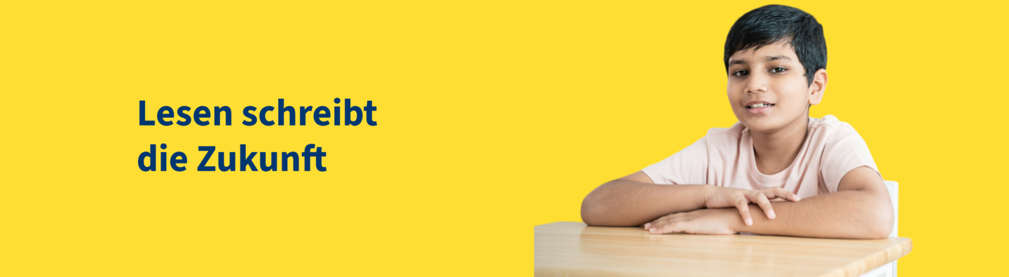 Ein Junge sitzt an einem Tisch vor gelbem Hintergrund und lächelt in die Kamera. Links daneben steht der Schriftzug „Lesen schreibt die Zukunft“ in blauer Schrift.