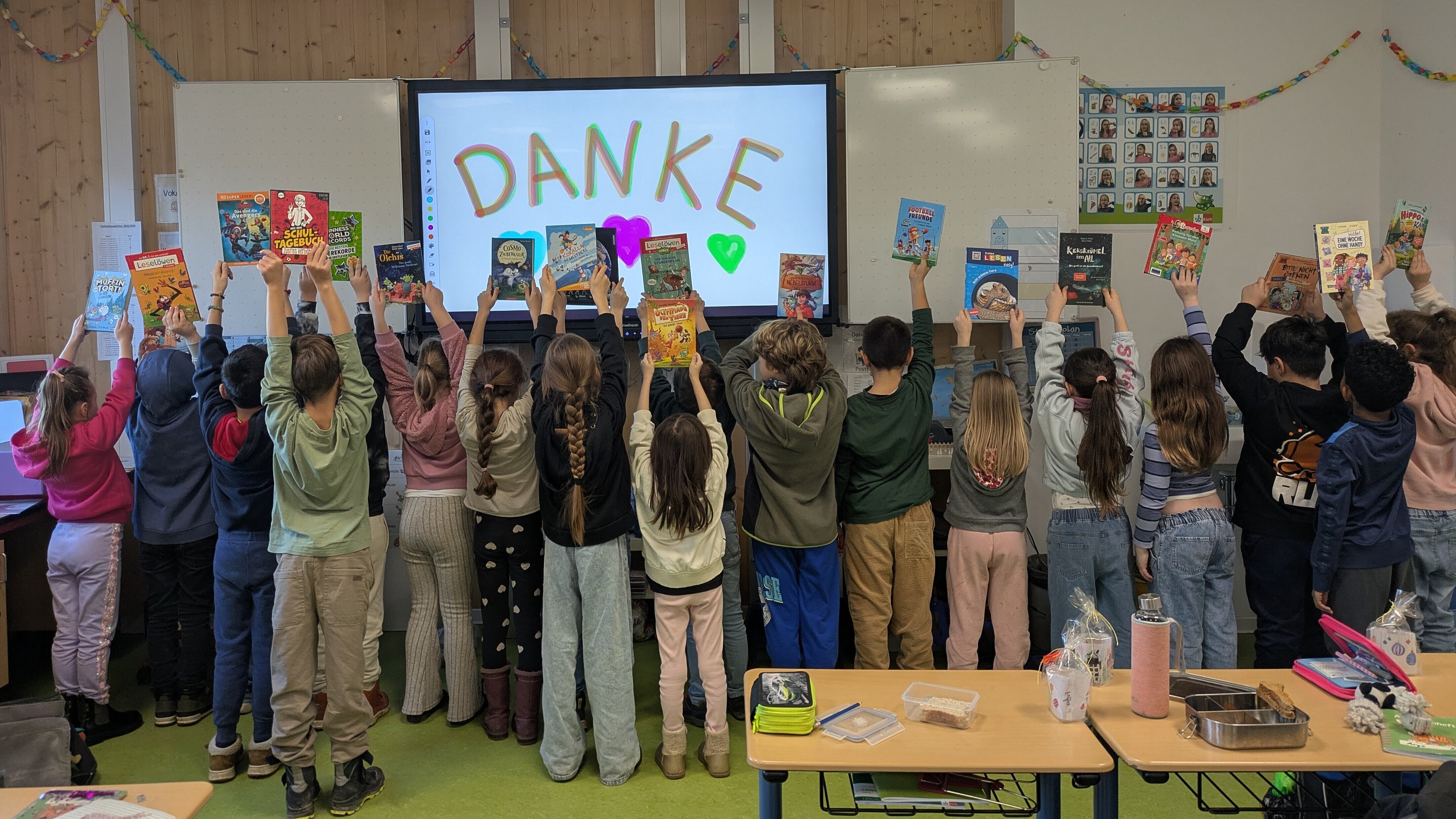 Eine Schulklasse steht mit dem Rücken zur Kamera im Klassenraum. Die Kinder halten Bücher über ihre Köpfe; auf dem Smartboard im Hintergrund steht groß „DANKE“ mit bunten Herzen.
