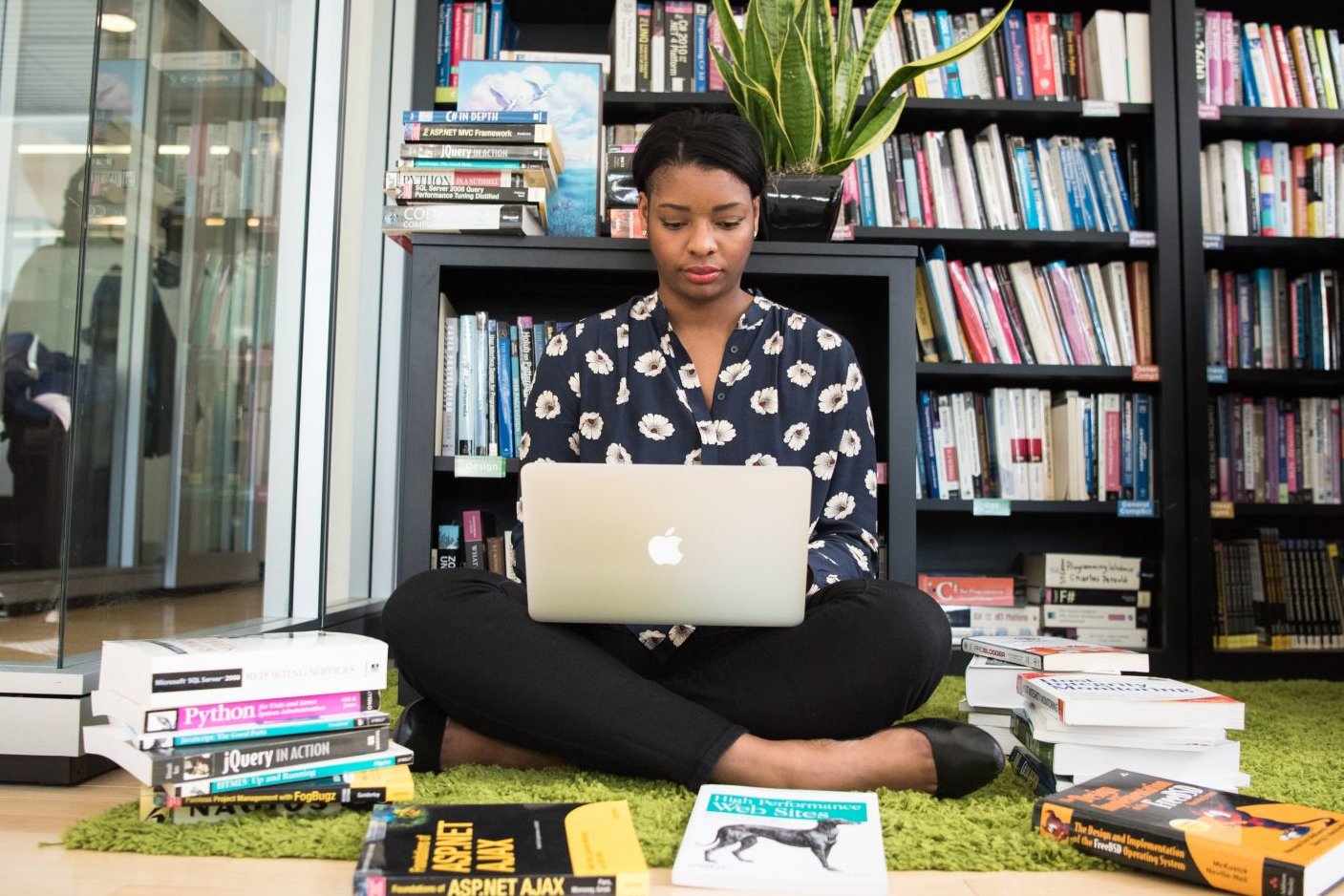 Junge Frau sitzt mit Laptop vor Bücherregal – vor ihr auf dem Boden liegen Bücher