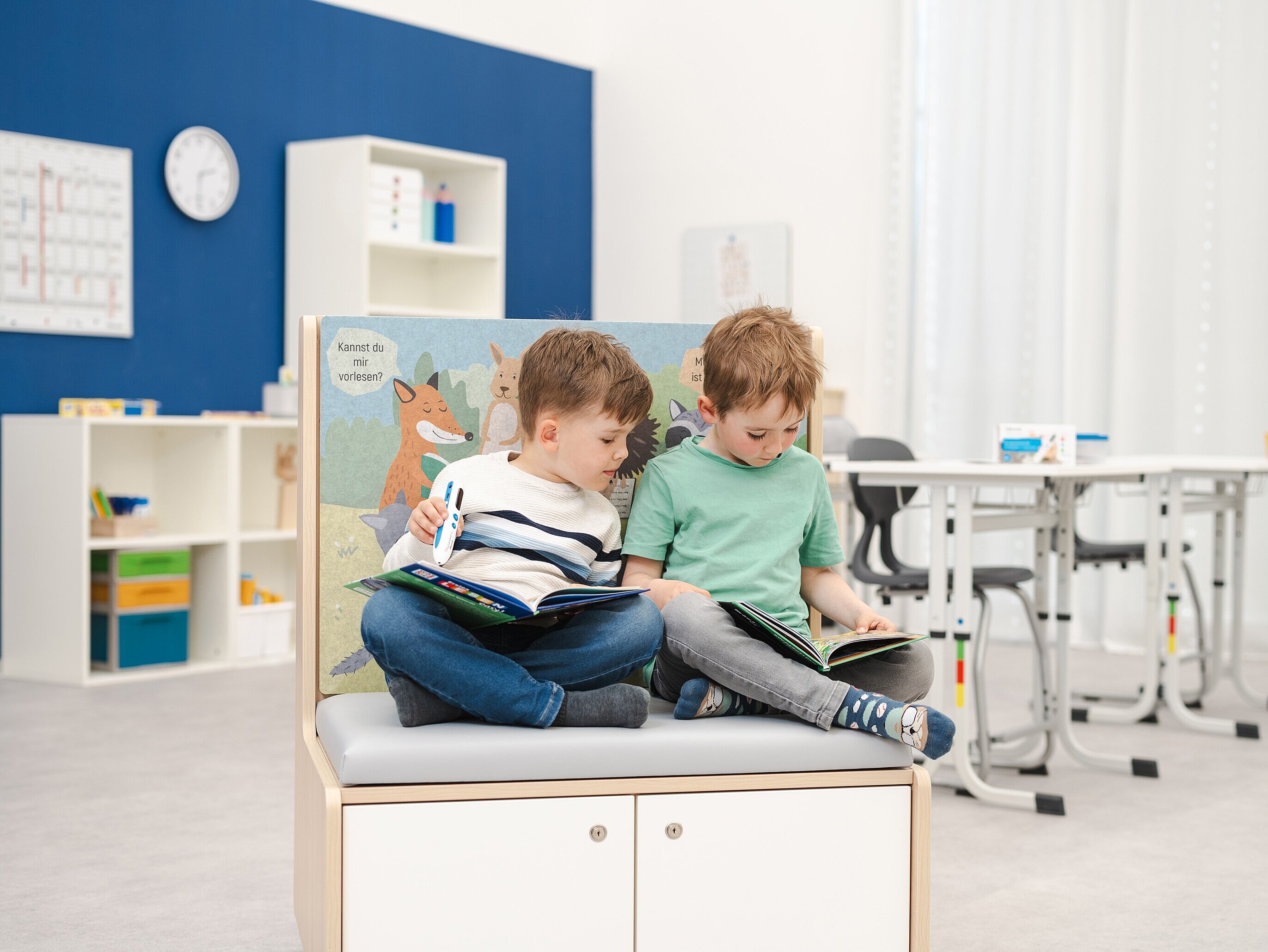 Zwei Kinder sitzen auf einem Lese-Möbel in einem hellen Klassenraum und schauen in Bilderbücher.