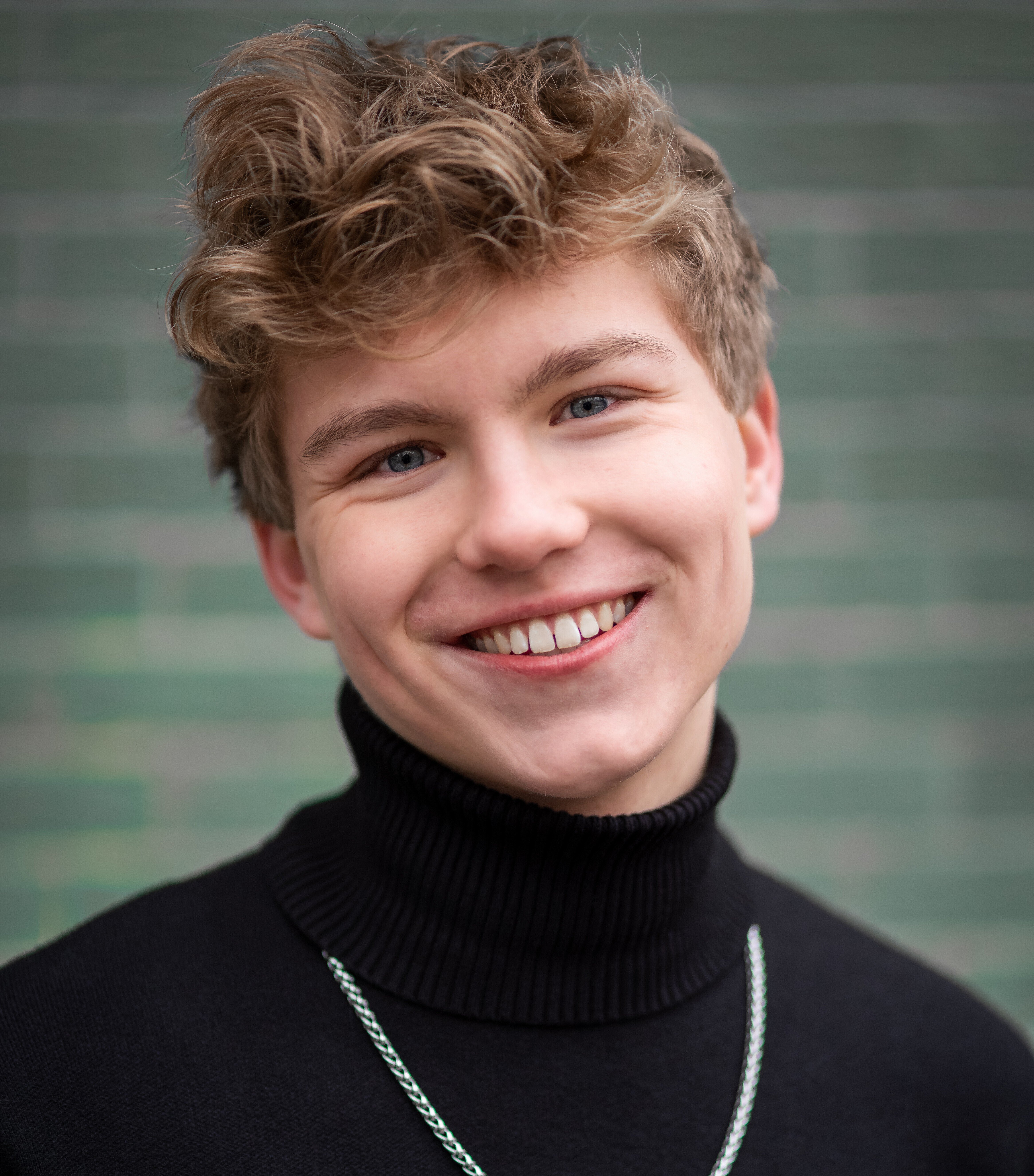 Porträt eines lächelnden Jugendlichen mit kurzen, lockigen Haaren, schwarzem Rollkragenpullover und Kette vor einem unscharfen, grünen Hintergrund.