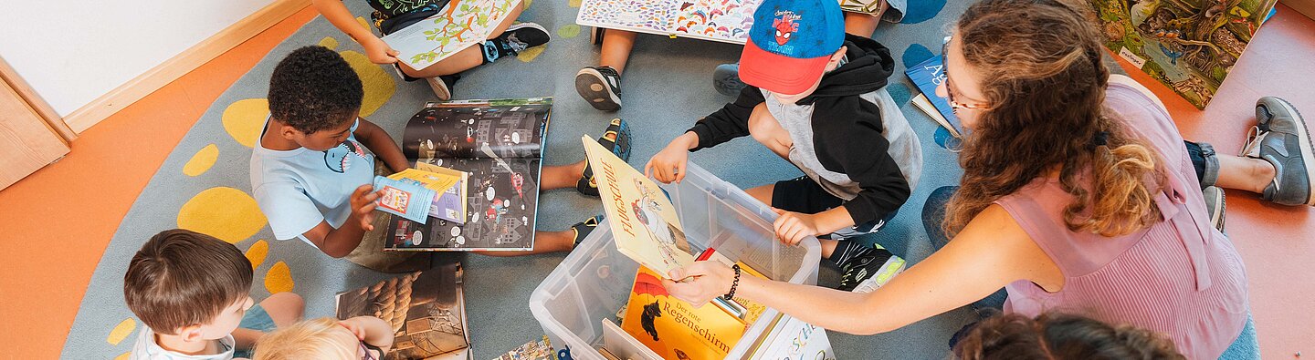 Mehrere Kinder sitzen mit zwei betreuenden Erwachsenen im Kreis auf einem bunten Teppich in einer Kita und entdecken gemeinsam Bilderbücher, die aus großen Boxen in der Mitte ausgewählt werden.