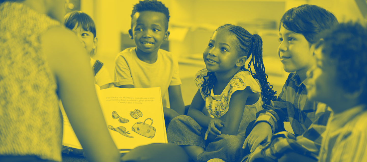 Mehrere Kinder sitzen zusammen und hören aufmerksam einer vorlesenden Person zu, während sie ein Bilderbuch betrachten.