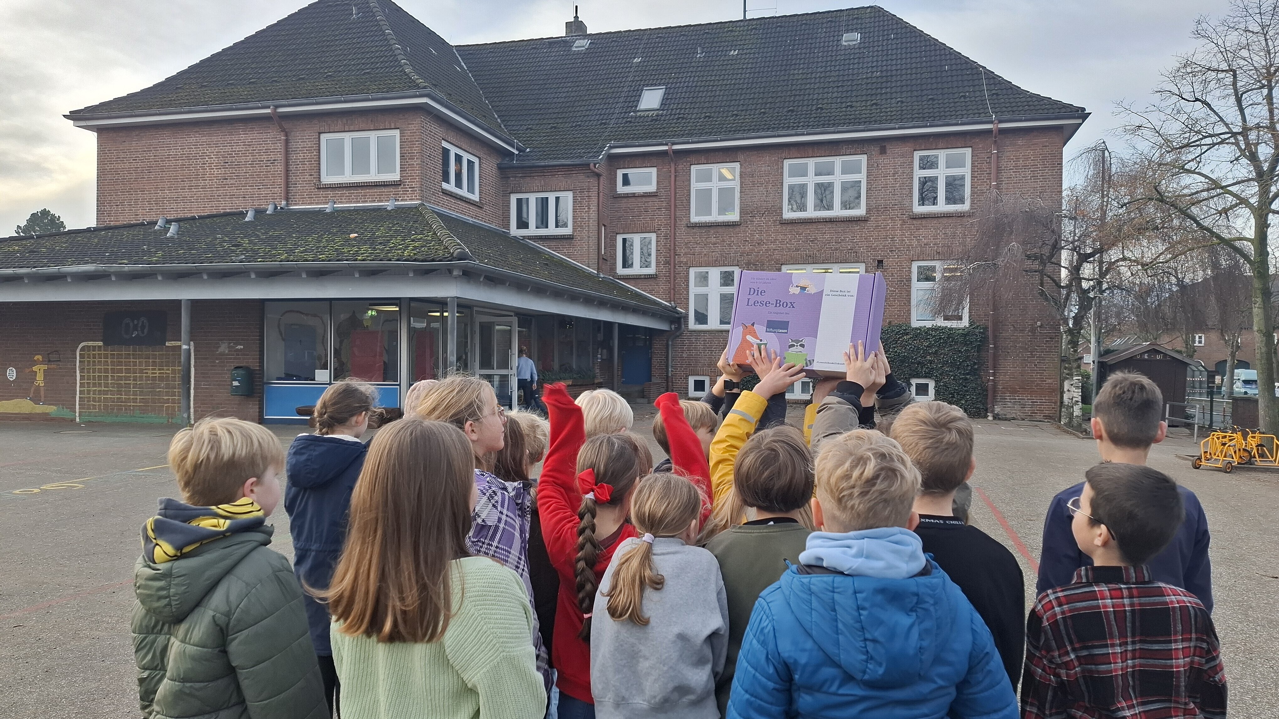 Eine Gruppe von Schulkindern steht auf einem Schulhof vor einem Backsteingebäude. Mehrere Kinder halten gemeinsam eine violette „Lese-Box“ über ihre Köpfe.