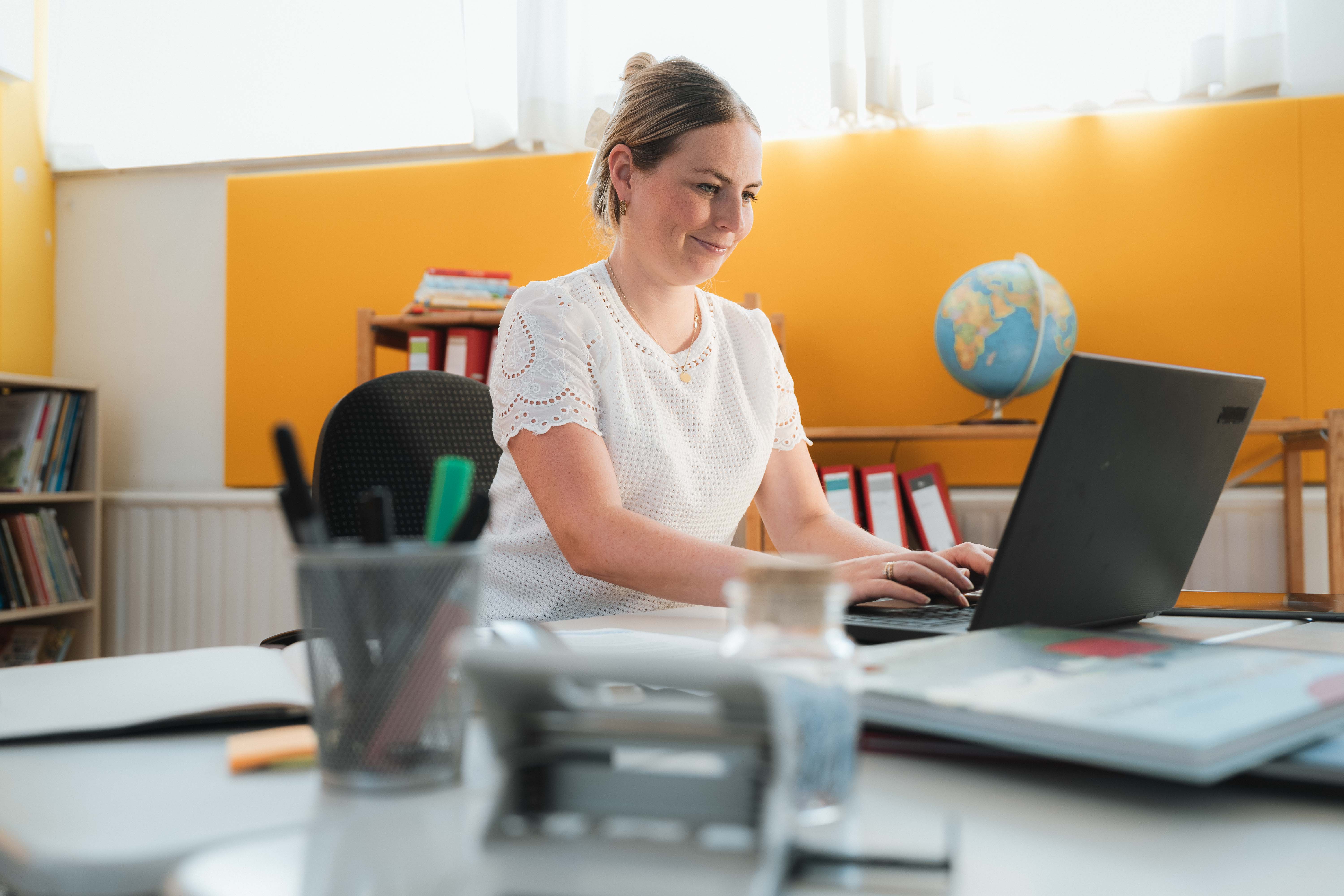 Eine Person sitzt in einem hellen Arbeitsraum an einem Schreibtisch und arbeitet auf einem Laptop, umgeben von Büromaterial, Ordnern und einer Weltkugel im Hintergrund.